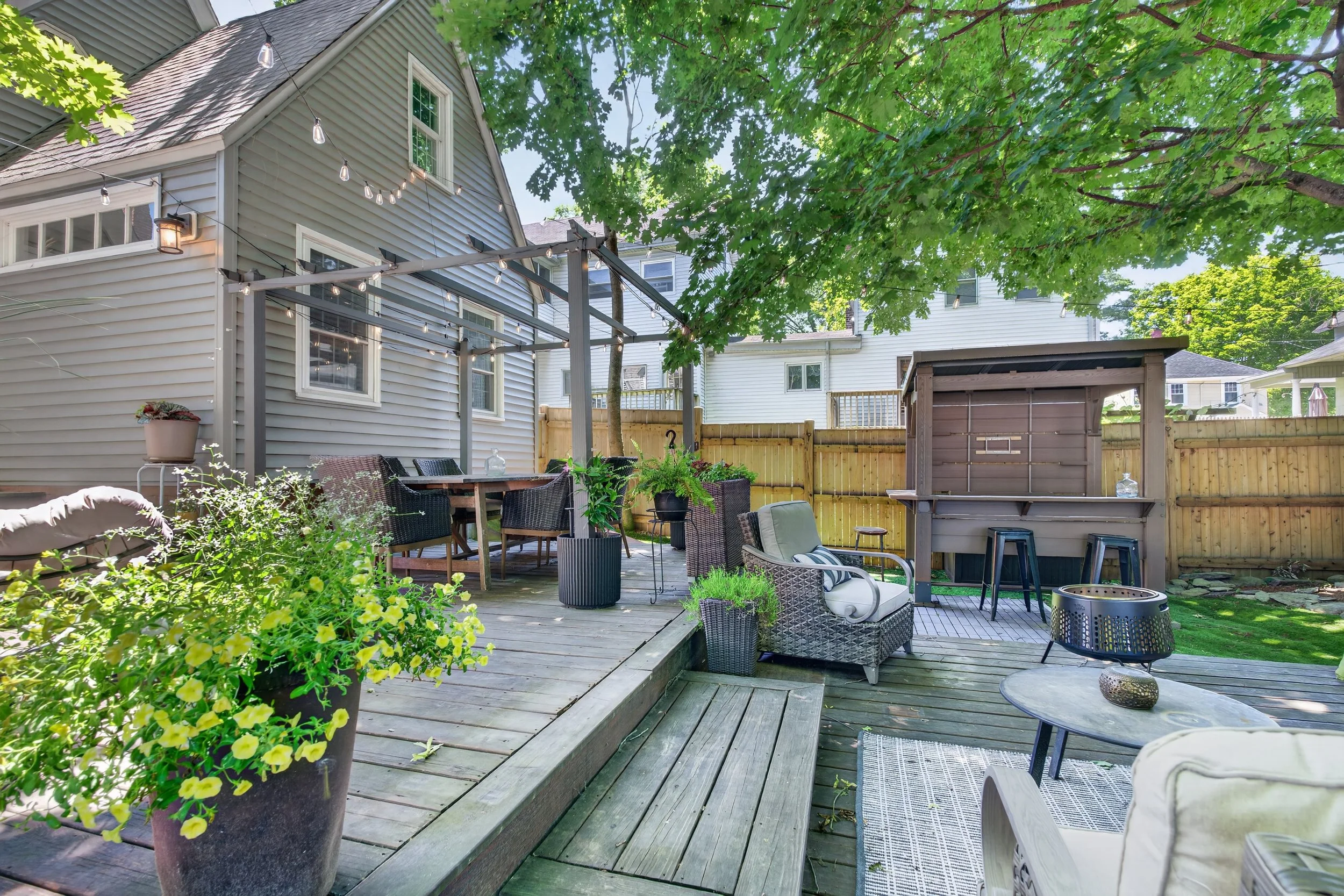 A backyard deck area with outdoor furniture, potted plants, string lights, and a wooden shed, surrounded by green trees and neighboring houses.