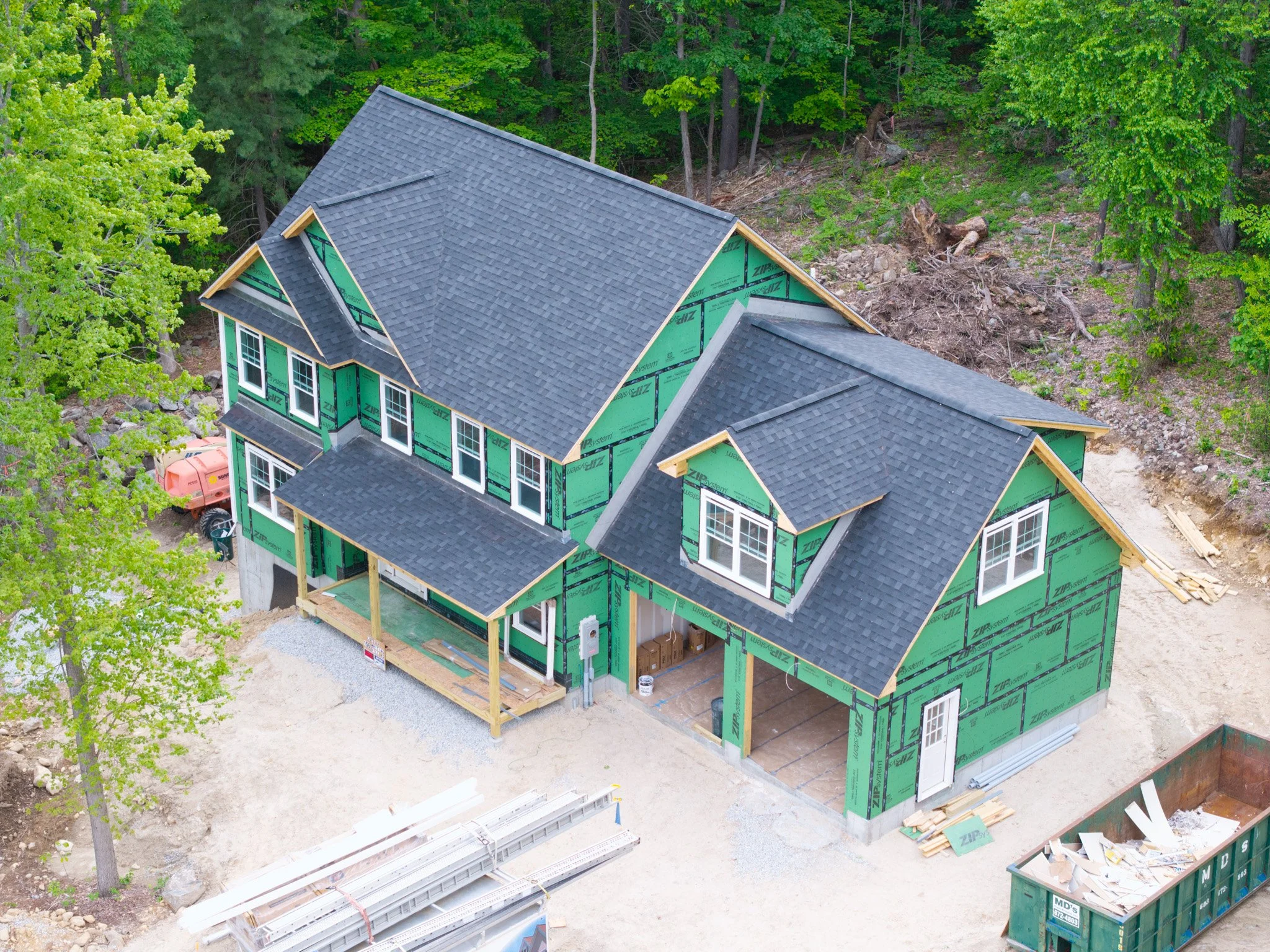 A house under construction in a wooded area, with green sheathing and a completed gray shingle roof. Construction materials and equipment are visible around the house.