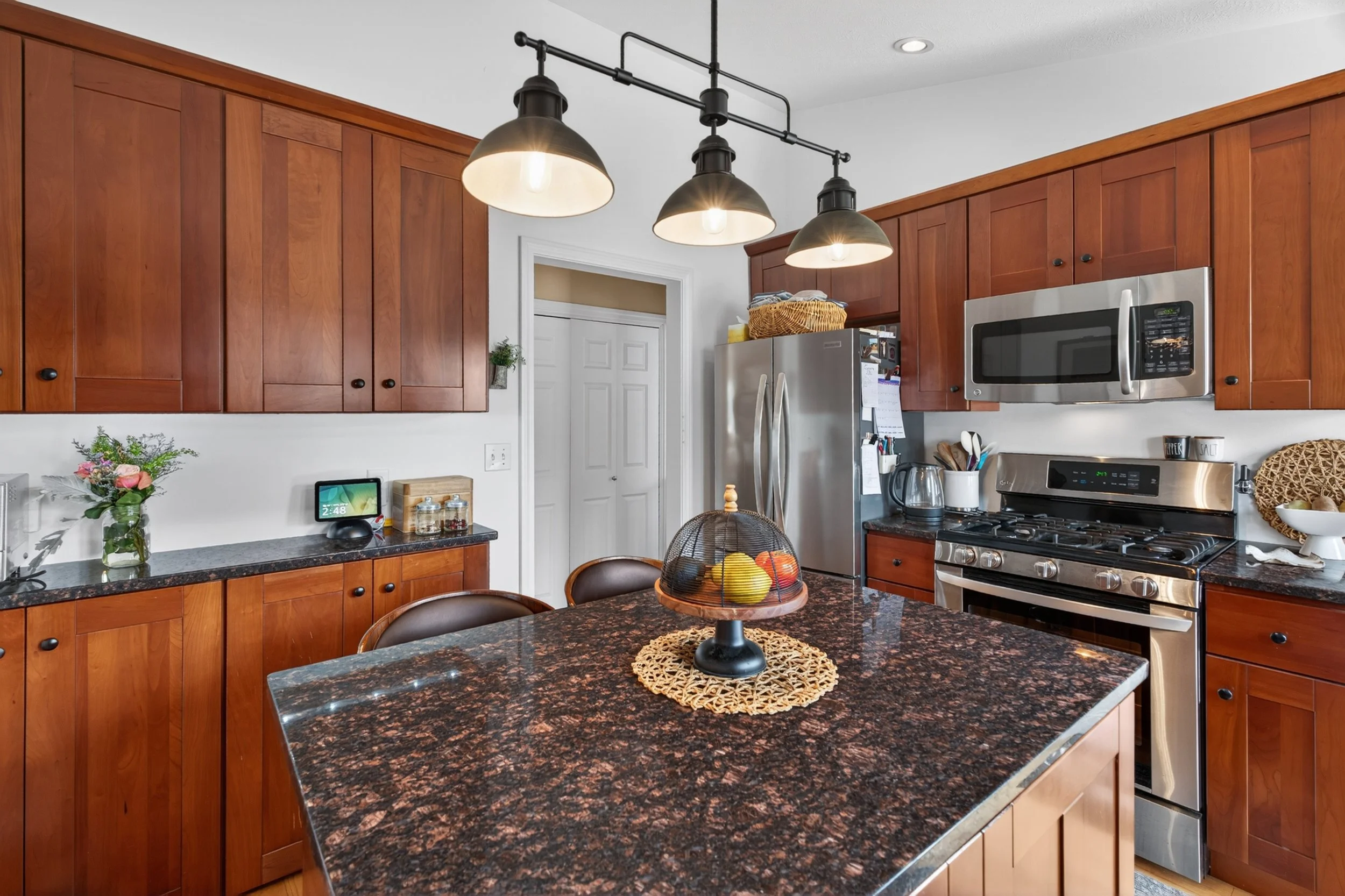 A kitchen with wooden cabinets, a granite island, stainless steel refrigerator, oven, microwave, and colorful fruit basket. There are decorative flowers, a small touchscreen device, and a jar on the countertops.