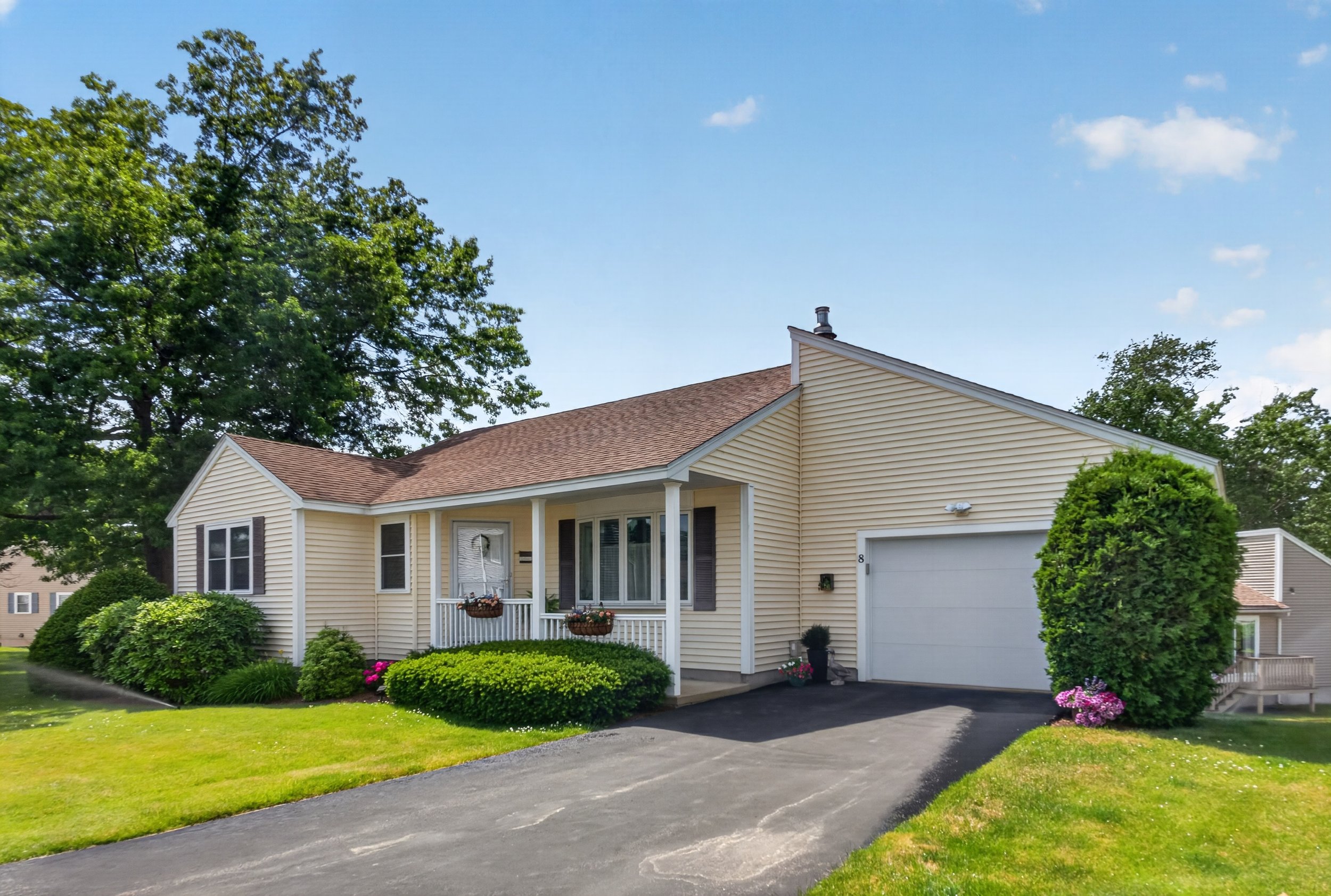 A yellow house with a brown shingle roof, front porch with hanging flower baskets, white railing, and a white garage door, surrounded by well-maintained green lawn and bushes, under a clear blue sky.