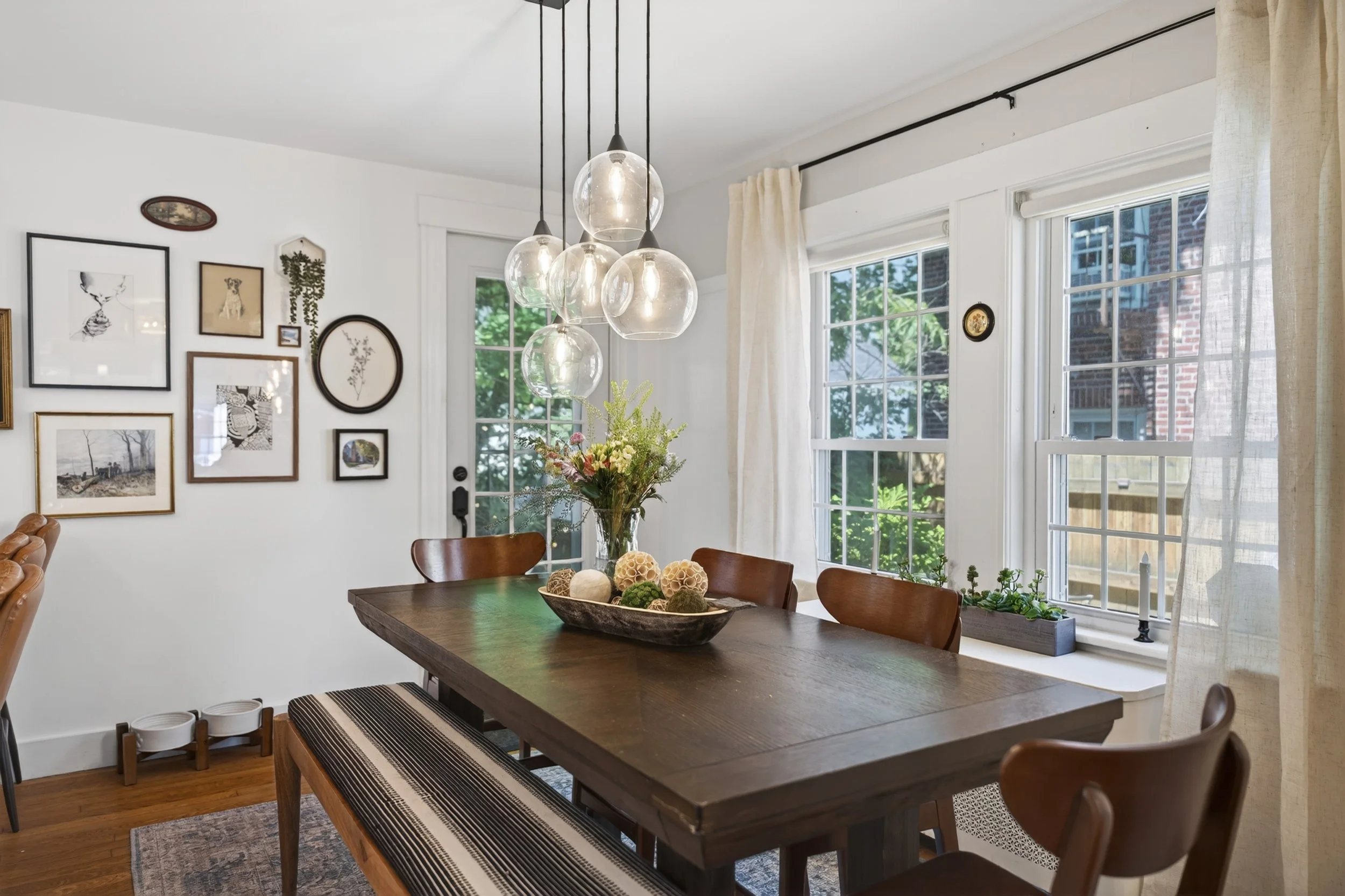 A dining room with a wooden table, surrounded by chairs, a hanging light fixture with multiple glass globes, and large windows with white curtains. The wall features a collection of framed artwork and a small hanging plant, with trees visible outside