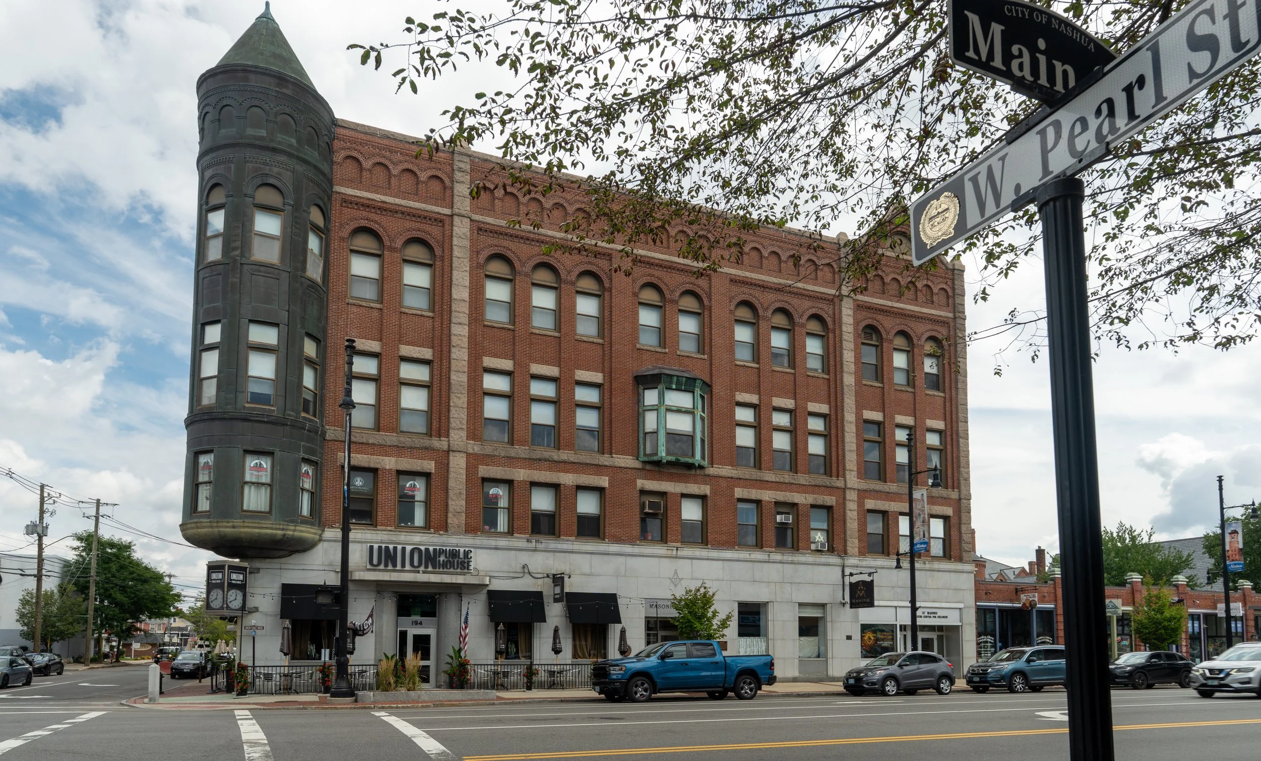 A multi-story brick building with arched windows and a turret on the corner, labeled 'Union Public House' on the lower level. Street signs indicate the intersection of Main Street and W. Pearl Street in Nashua. There are cars parked in front, and the