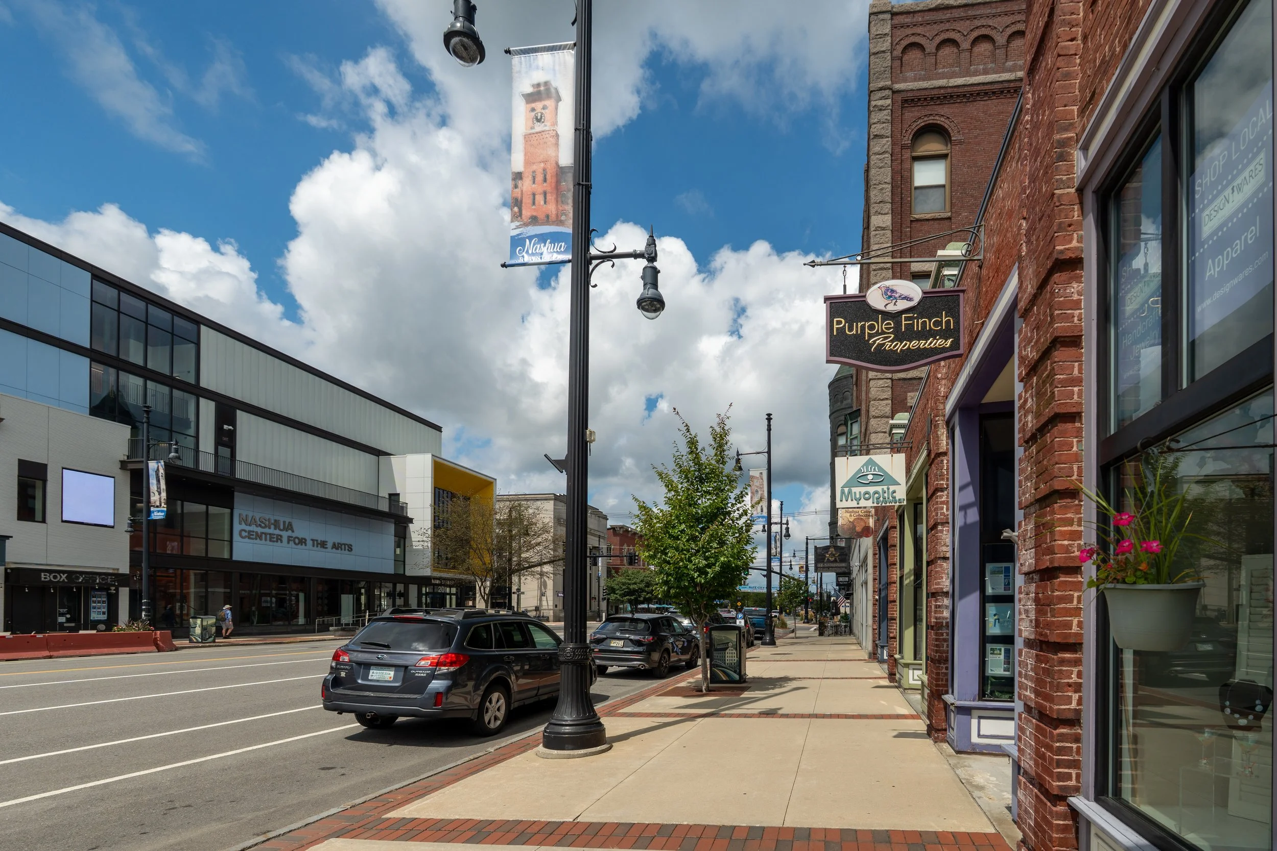 A city street with parked cars, a brick building with a sign that reads 'Purple Finch Properties,' and a modern glass building across the street. There is a lamppost with banners, one that says 'Nashua,' and a cloudy blue sky overhead.