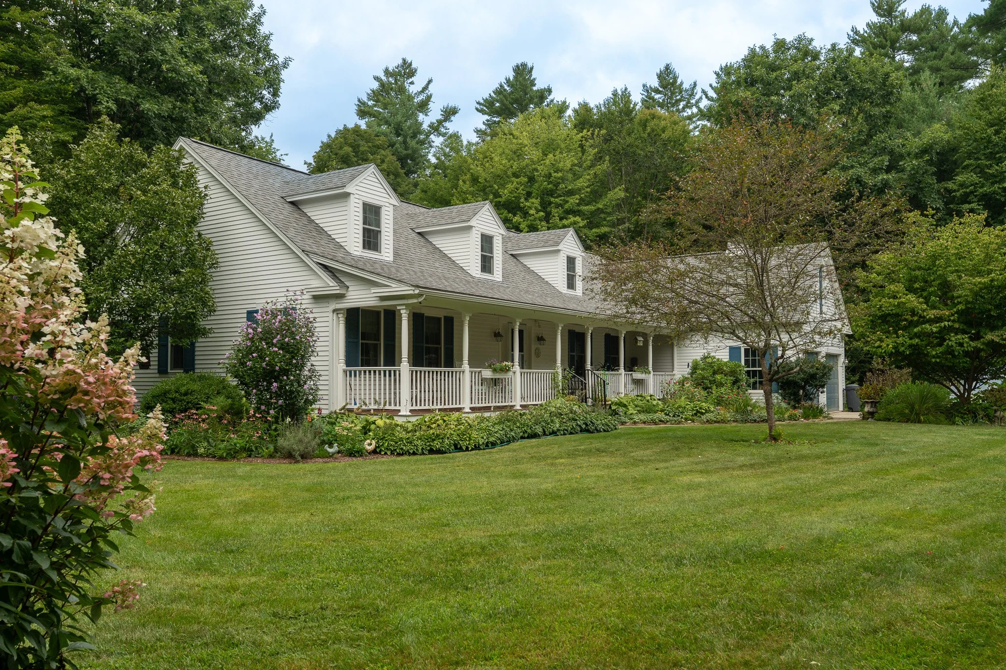 A white two-story house with a porch, surrounded by a lush green lawn and garden with various bushes and trees, under a partly cloudy sky.