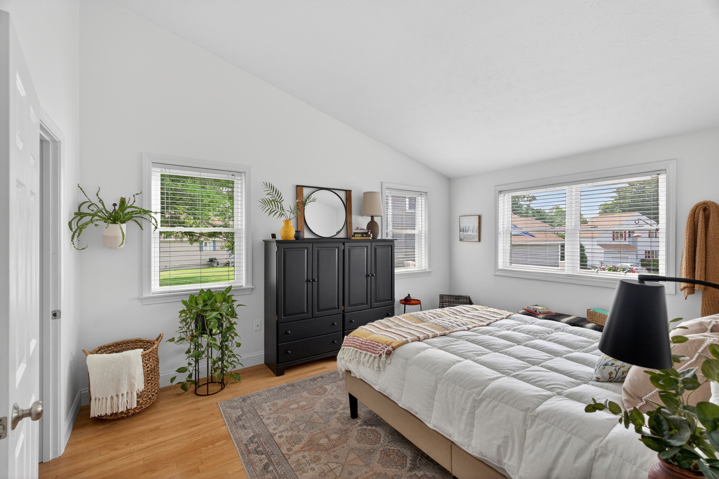 A bedroom with white walls, hardwood floors, and a sloped ceiling. The room features large windows with blinds, a black dresser with decorative vases and a mirror, a bed with a beige upholstery, a patterned blanket, and various plants and wicker furn