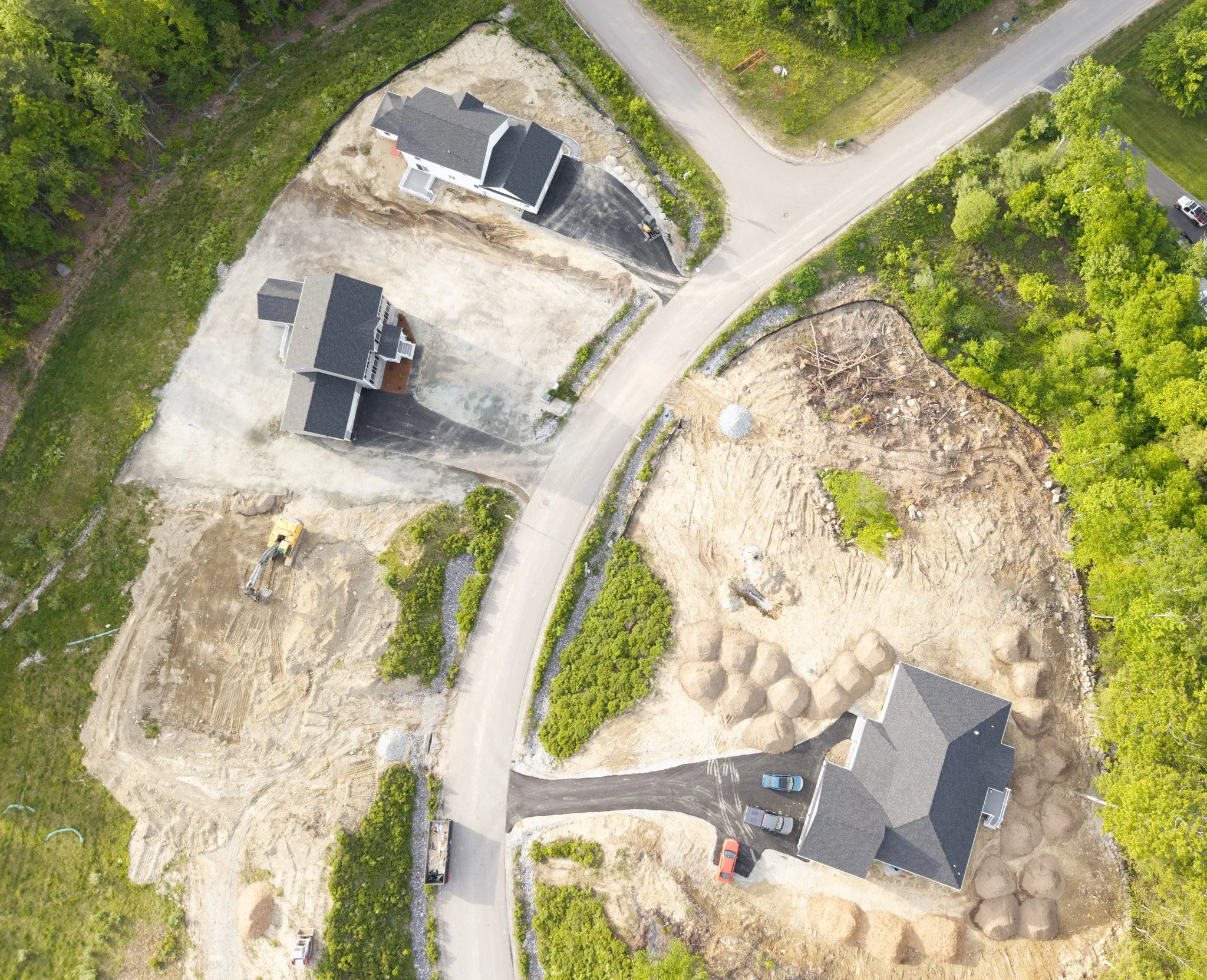 Aerial view of a residential construction site with partially built houses, dirt areas, sidewalks, and some parked cars surrounded by greenery.