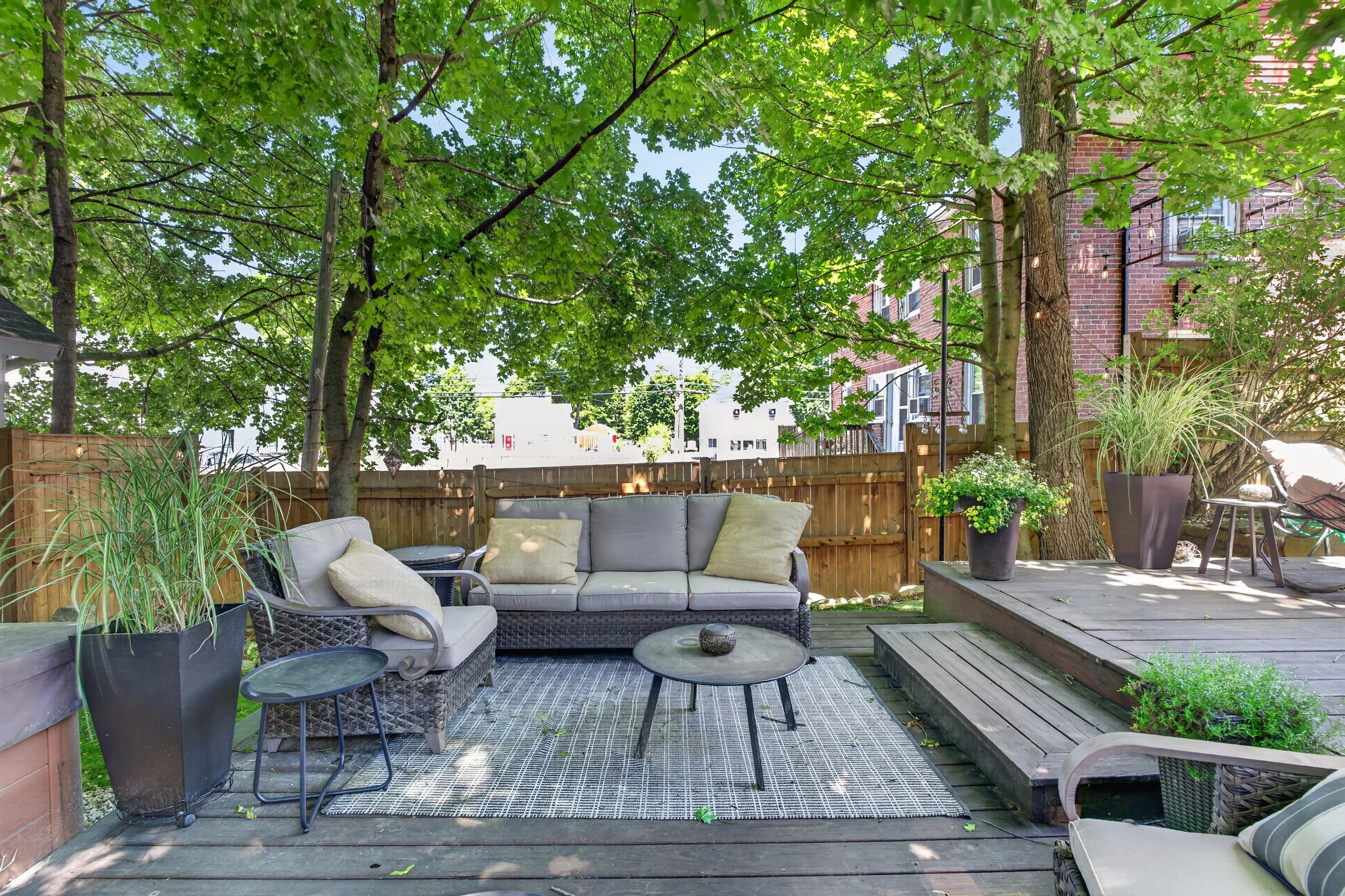 A backyard patio with outdoor seating under a canopy of green trees, with a wooden fence and brick buildings in the background.