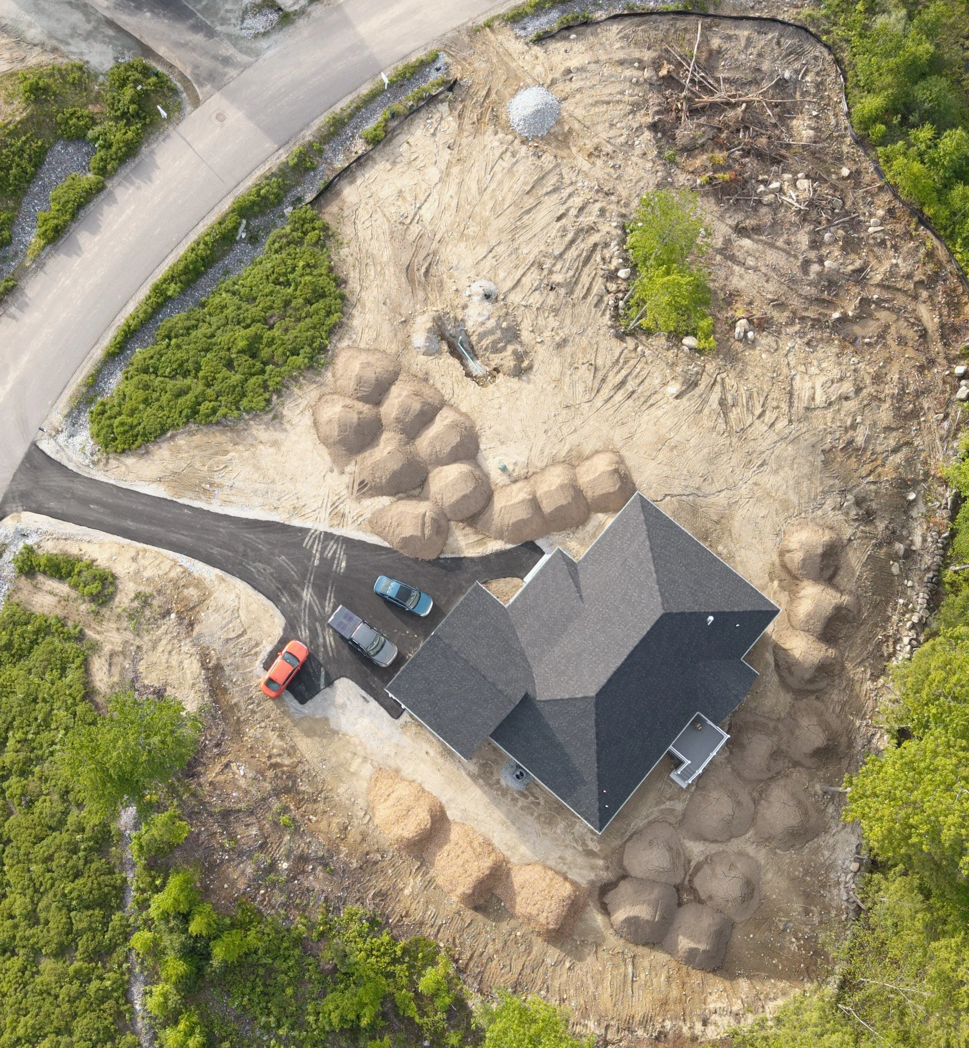 An aerial view of a construction site with cleared land, construction debris, and trees, adjacent to a paved road with parked cars.