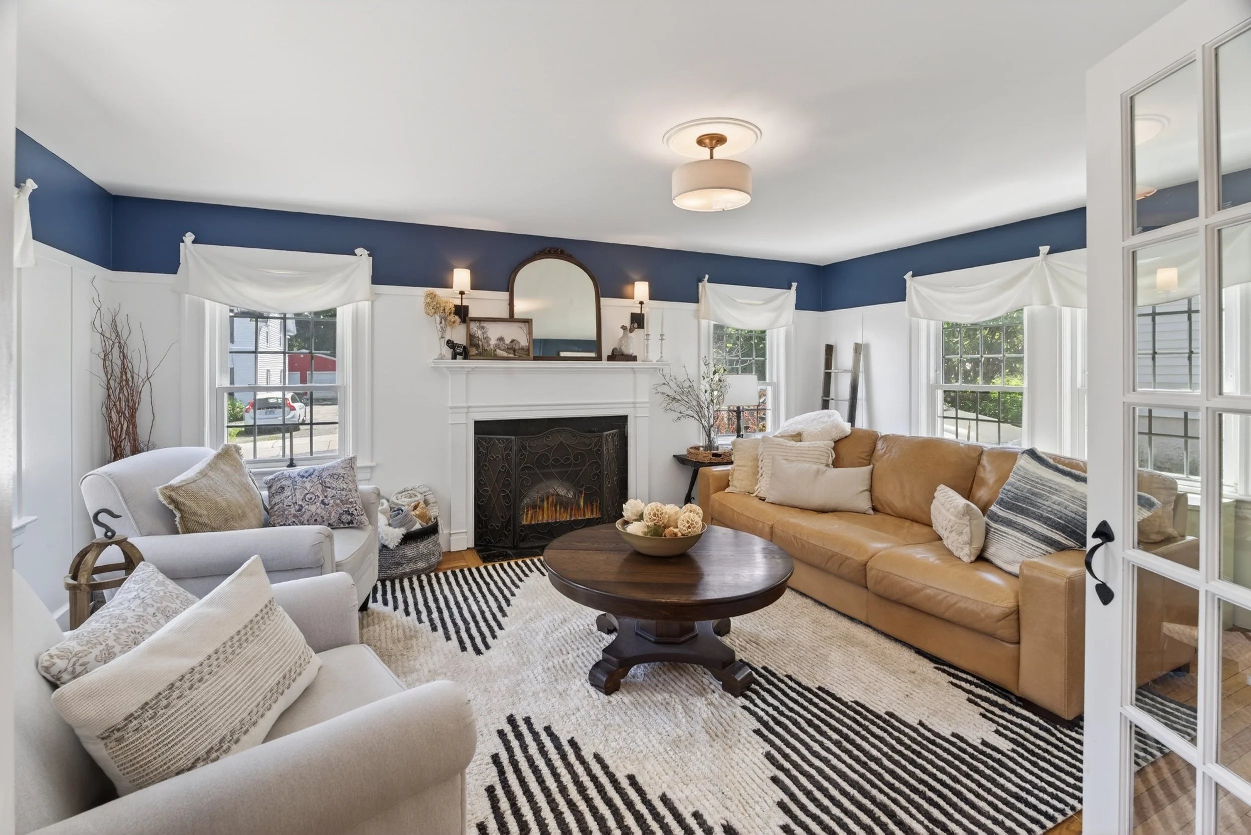 Living room with white and beige sofas, a round wooden coffee table, a fireplace, and multiple windows with white curtains. The room has a navy blue ceiling and light-colored walls, decorated with lamps, paintings, and decorative items.