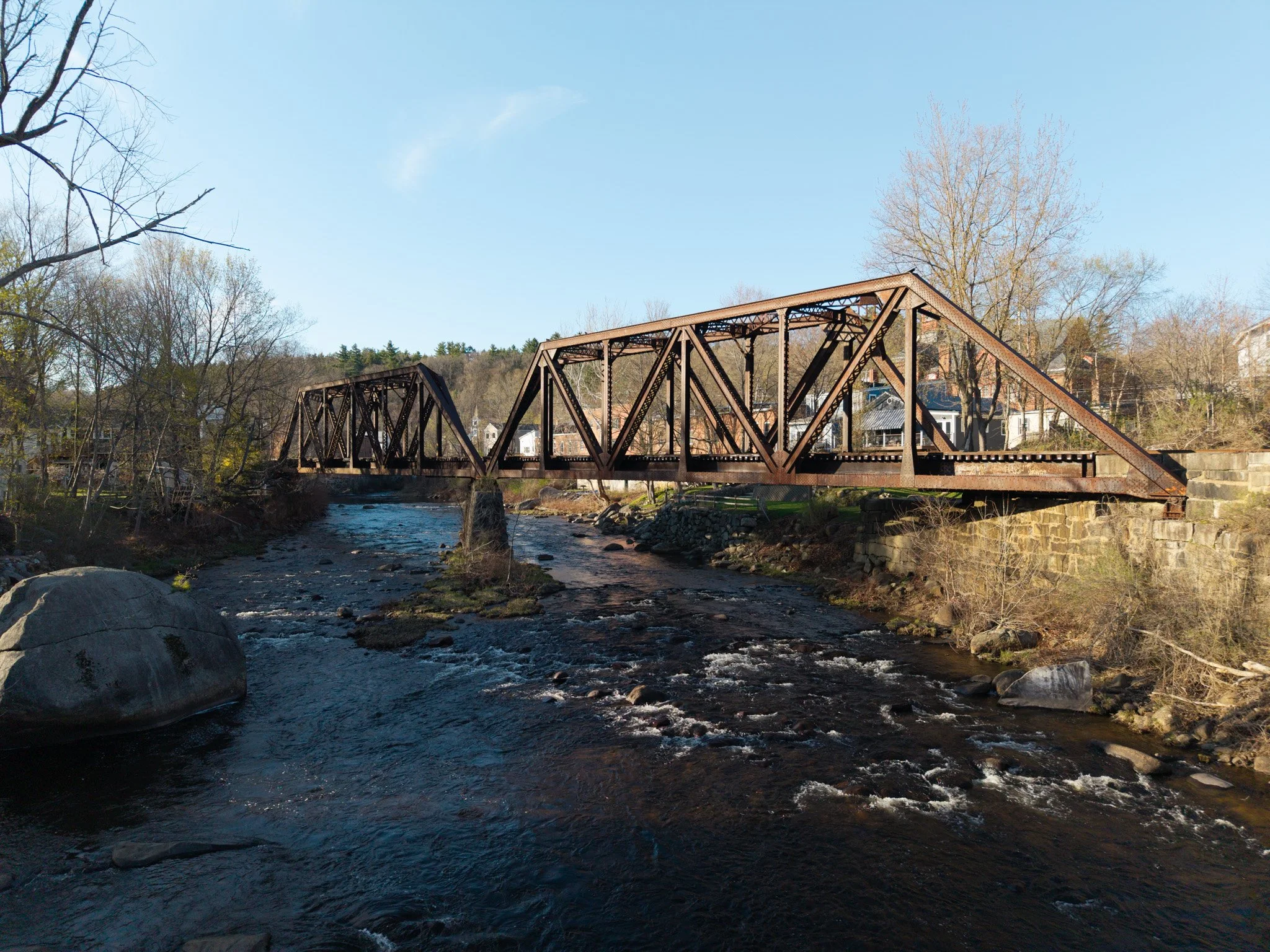 A rusted metal truss bridge crossing over a small river in a town, with trees, houses, and a hill in the background under a clear blue sky.