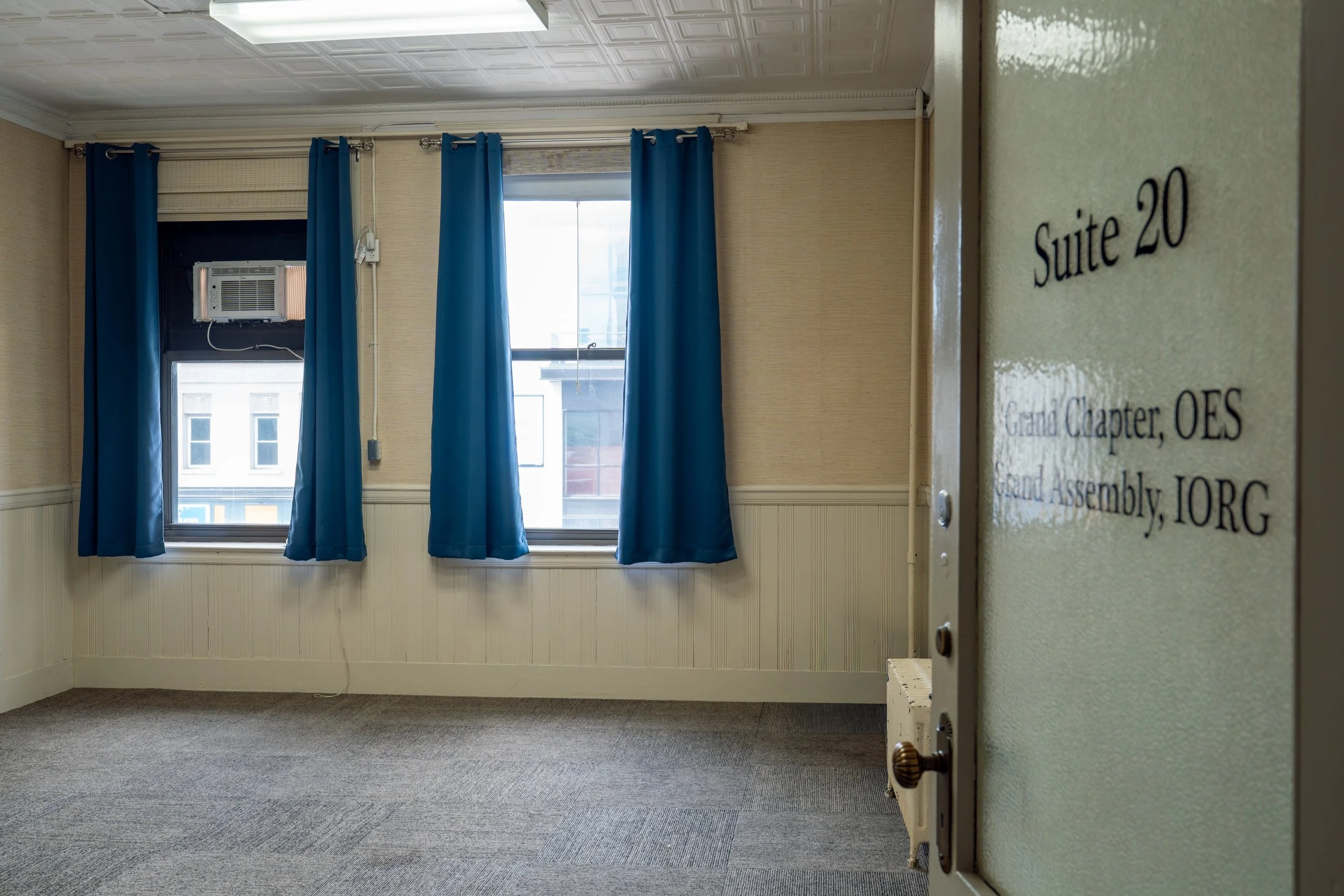 Empty room with beige walls, blue curtains on windows, gray carpet, and a small air conditioning unit in the window. A door with a sign reading 'Suite 20' is partly visible on the right.