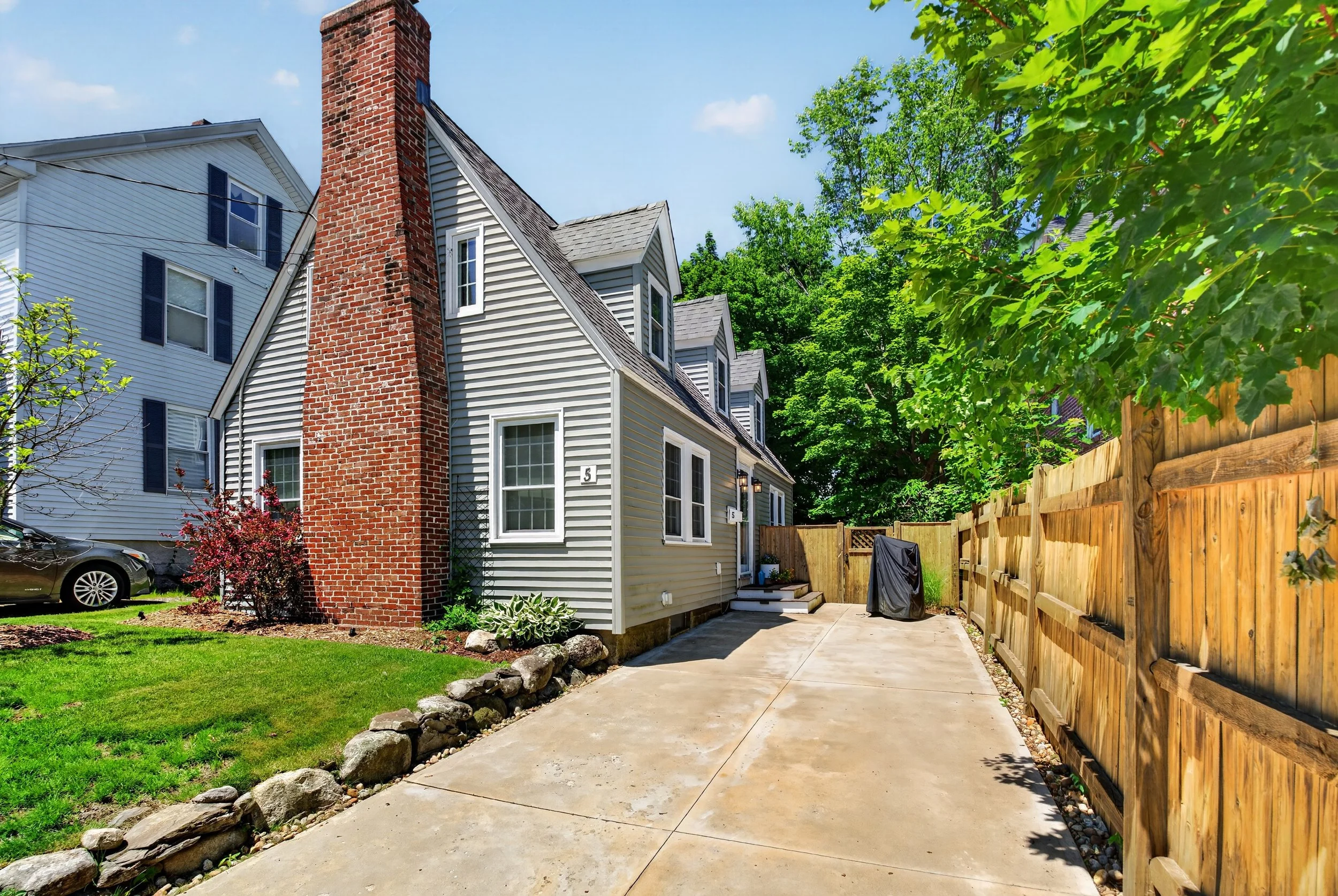 A backyard scene with a concrete patio, green lawn, a brick chimney attached to a white house with blue shutters, a wooden fence on the right, and lush green trees in the background under a blue sky.