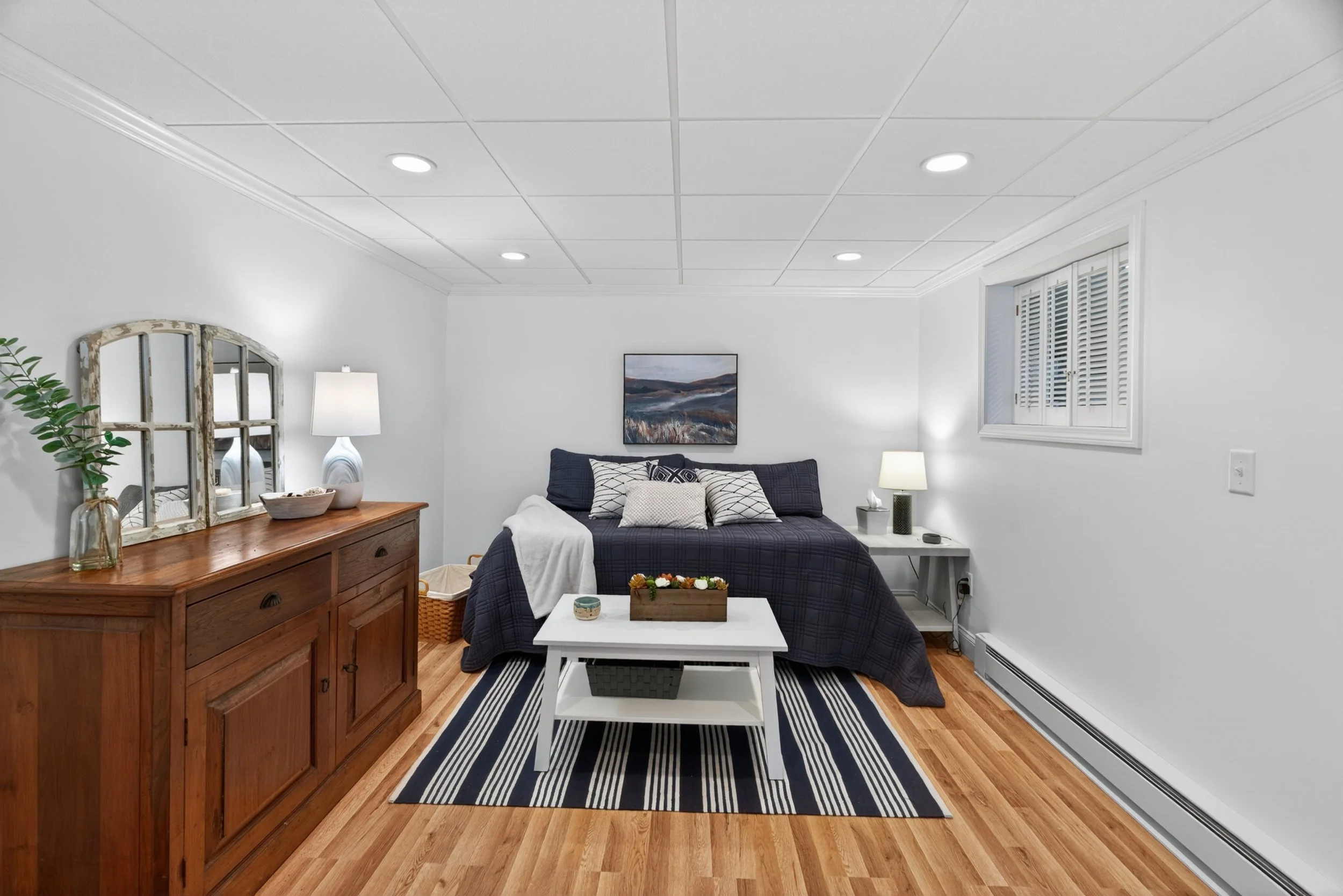Living room with white walls, wooden furniture, a black and white rug, a window with white shutters, and a bed with black bedding and multiple pillows, flanked by lamps.