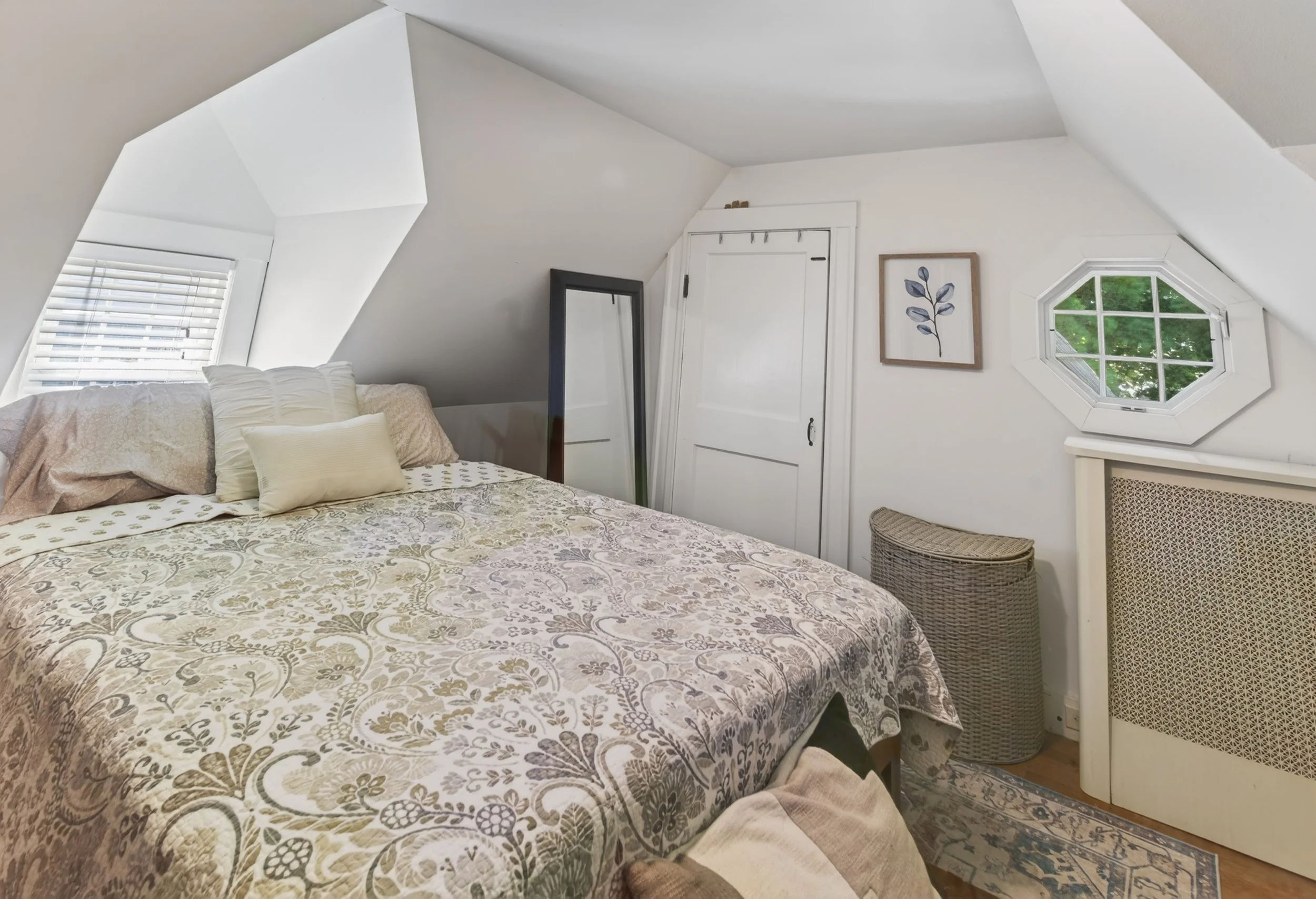 Photo of a cozy bedroom with a bed covered in a patterned quilt, pillows, a wicker basket, a mirror, a small framed botanical art, and a window with greenery outside, located in an attic with sloped ceilings.