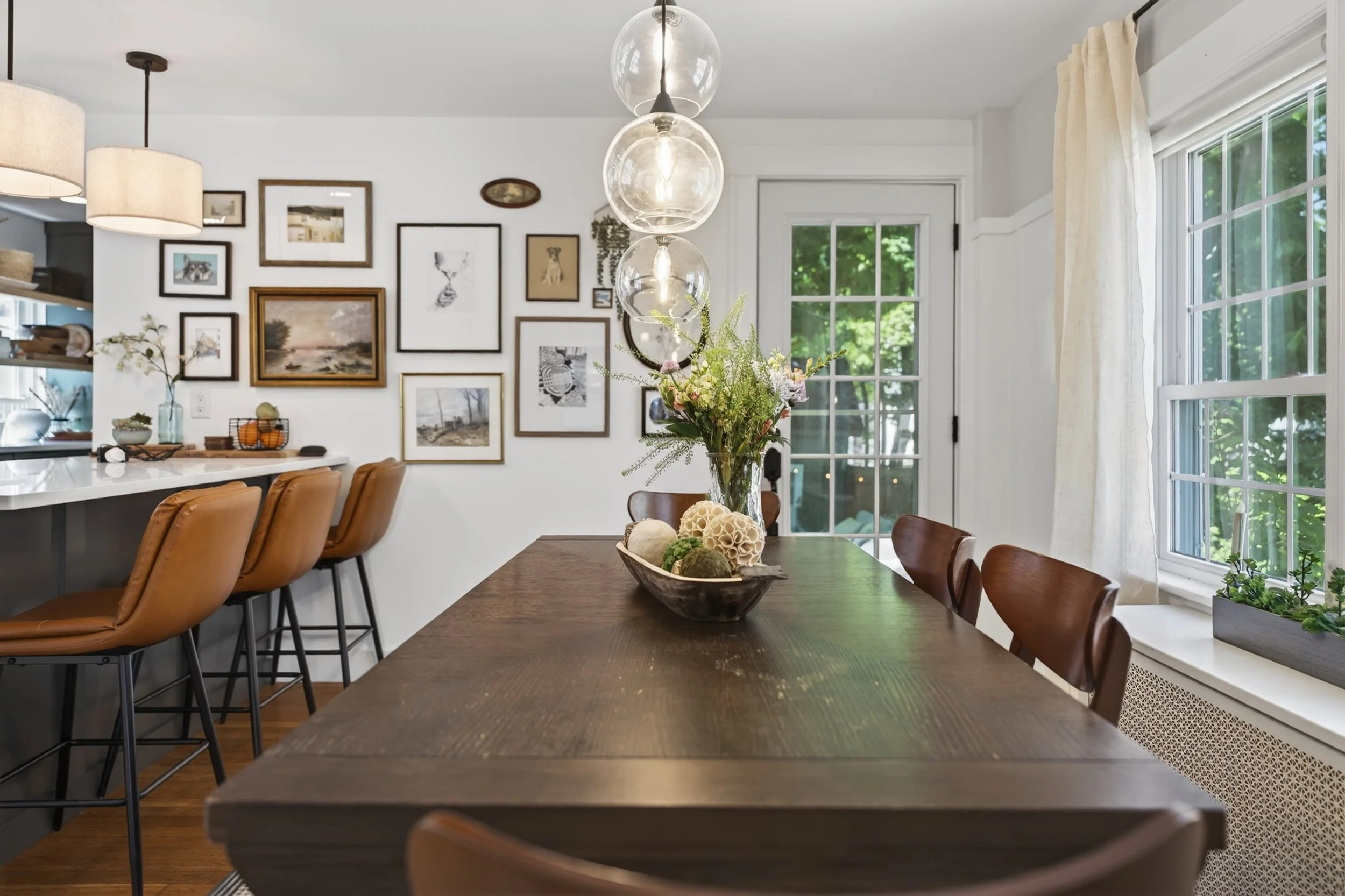 A dining room and kitchen area with a wooden dining table with chairs, decorative vases and flowers, framed artwork on the wall, large windows with curtains, and a hanging light fixture with three glass globes.