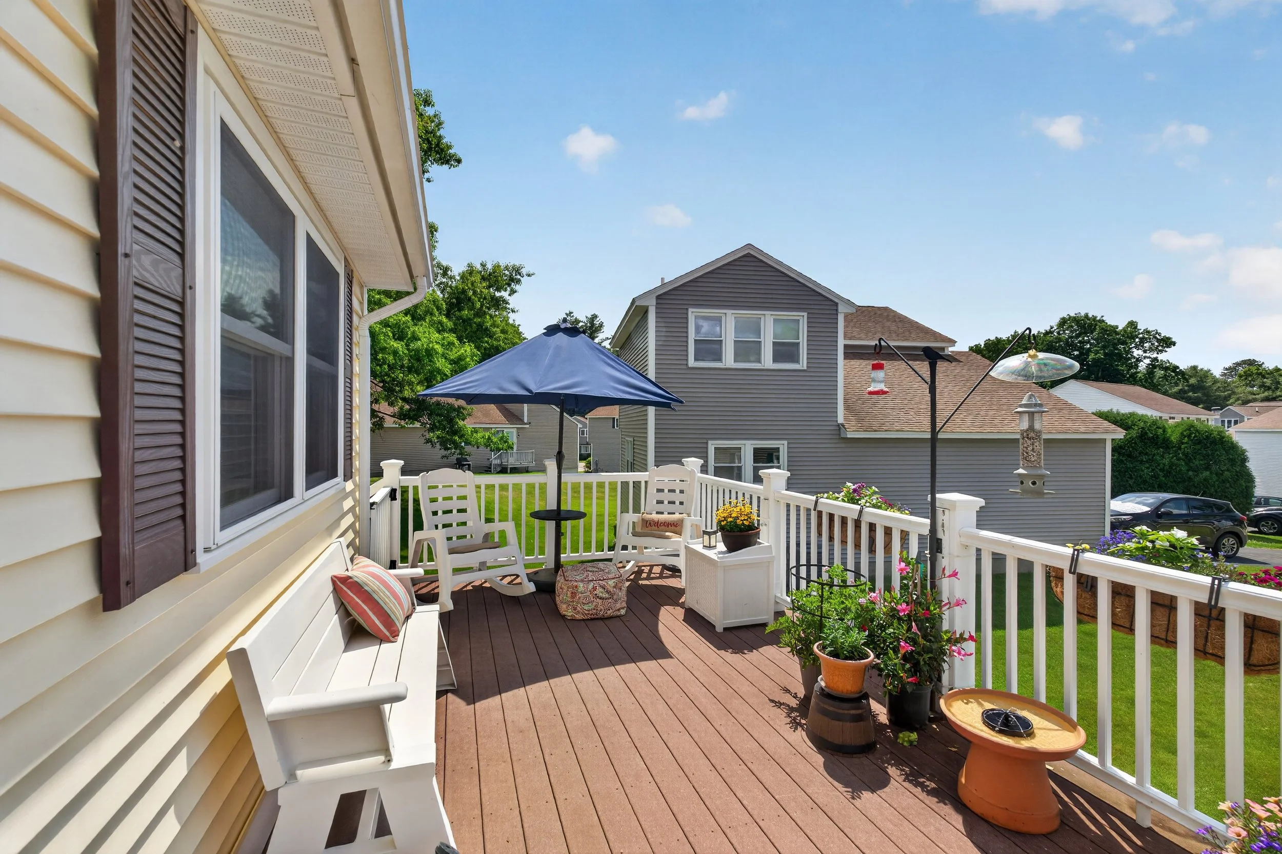 A backyard deck with white railing and outdoor furniture, including a bench with pillows, a small table, and white chairs under a blue umbrella. Potted plants with flowers decorate the deck. In the background, houses with gray and tan siding and shin