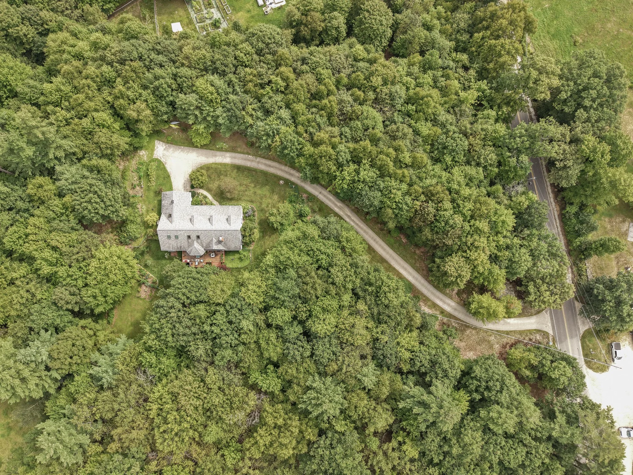An aerial view of a house surrounded by dense trees with a curved driveway, a road with a vehicle parked nearby, and some residential structures in the background.