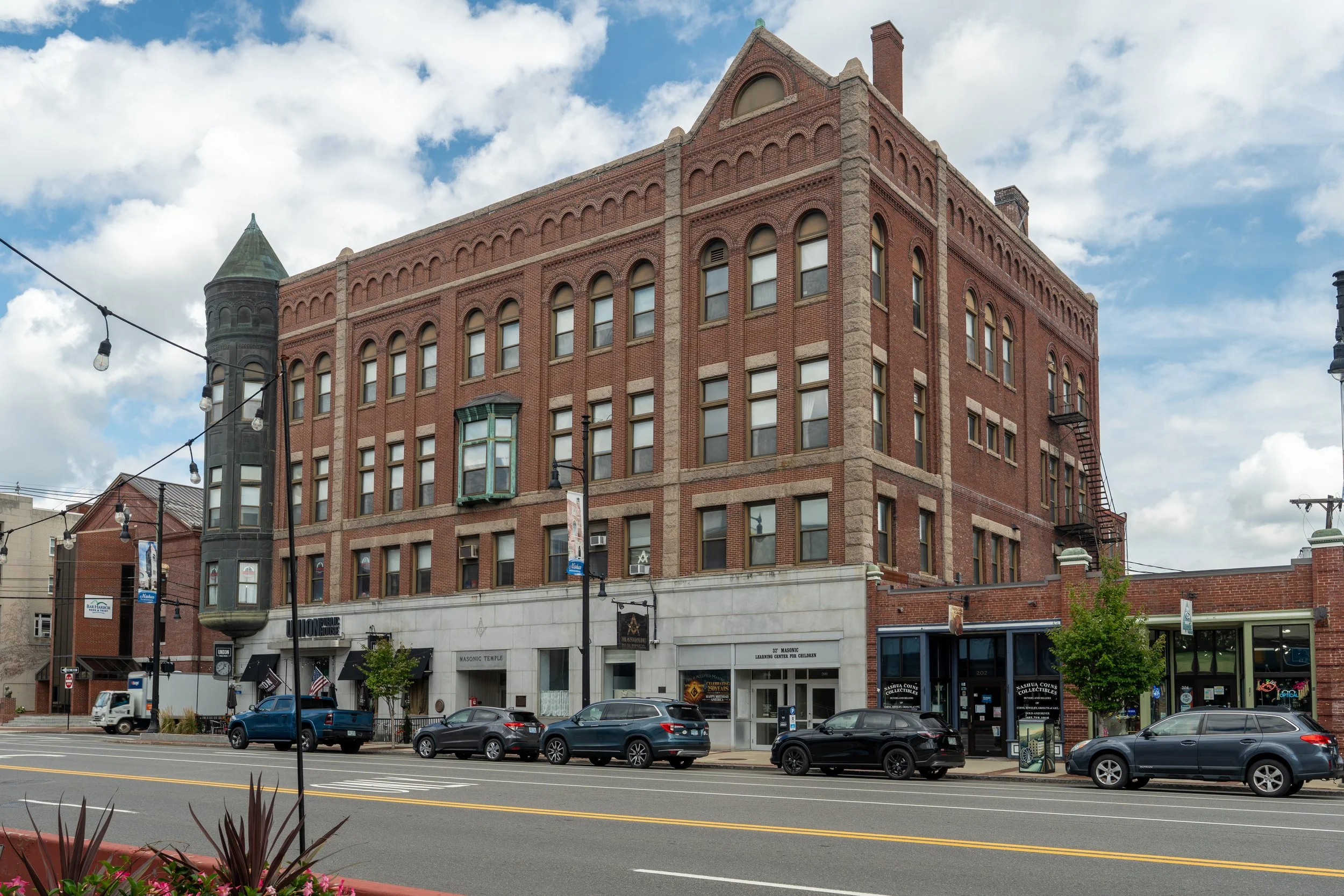 A historic red brick building with architectural details, large windows, and small towers at the corners, situated on a busy street with cars parked along the curb, string lights hanging across the street, and a blue sky with scattered clouds overhea