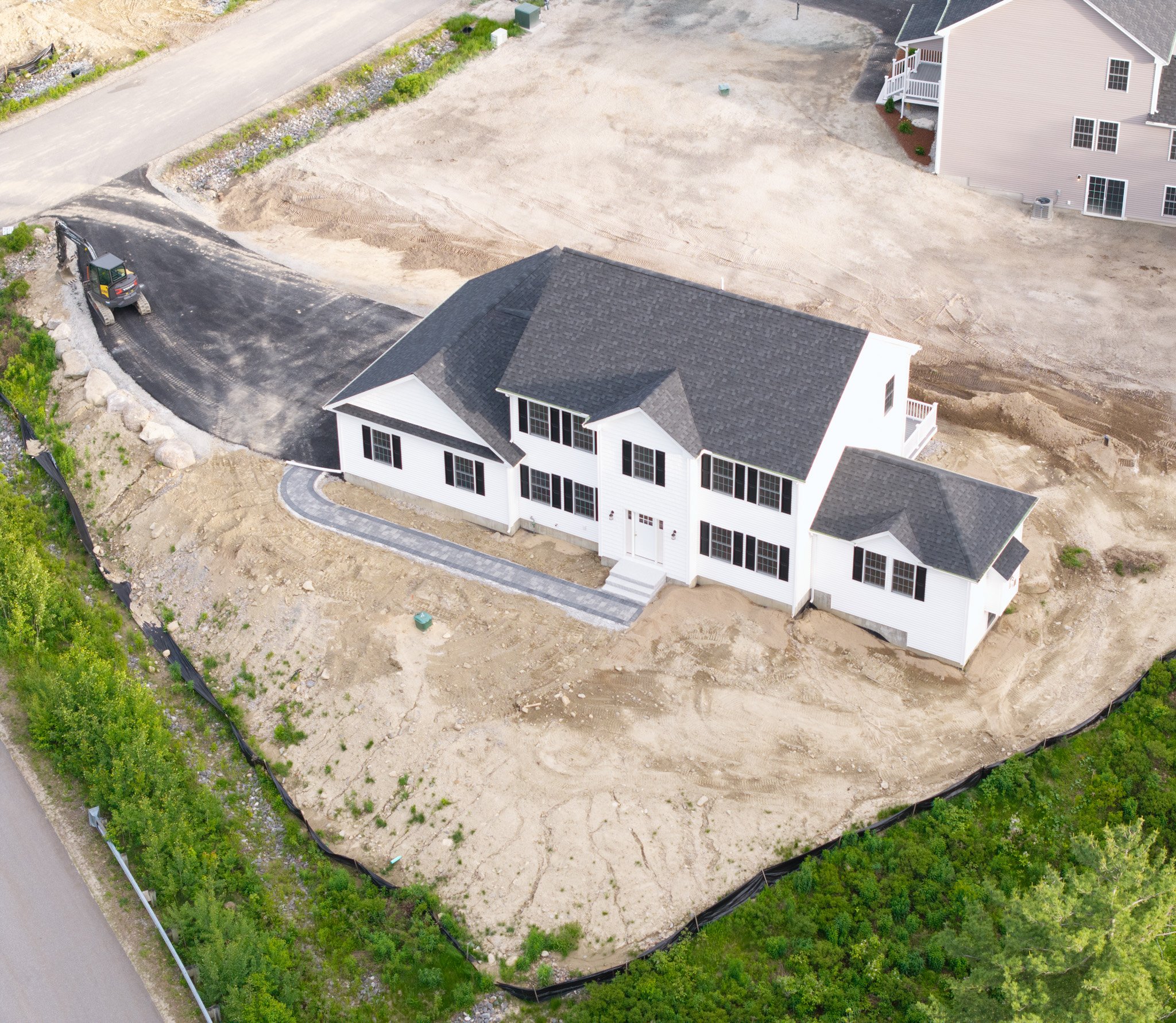 An aerial view of a residential area under construction with new houses, a cleared dirt lot, and a small excavator parked on a paved driveway.
