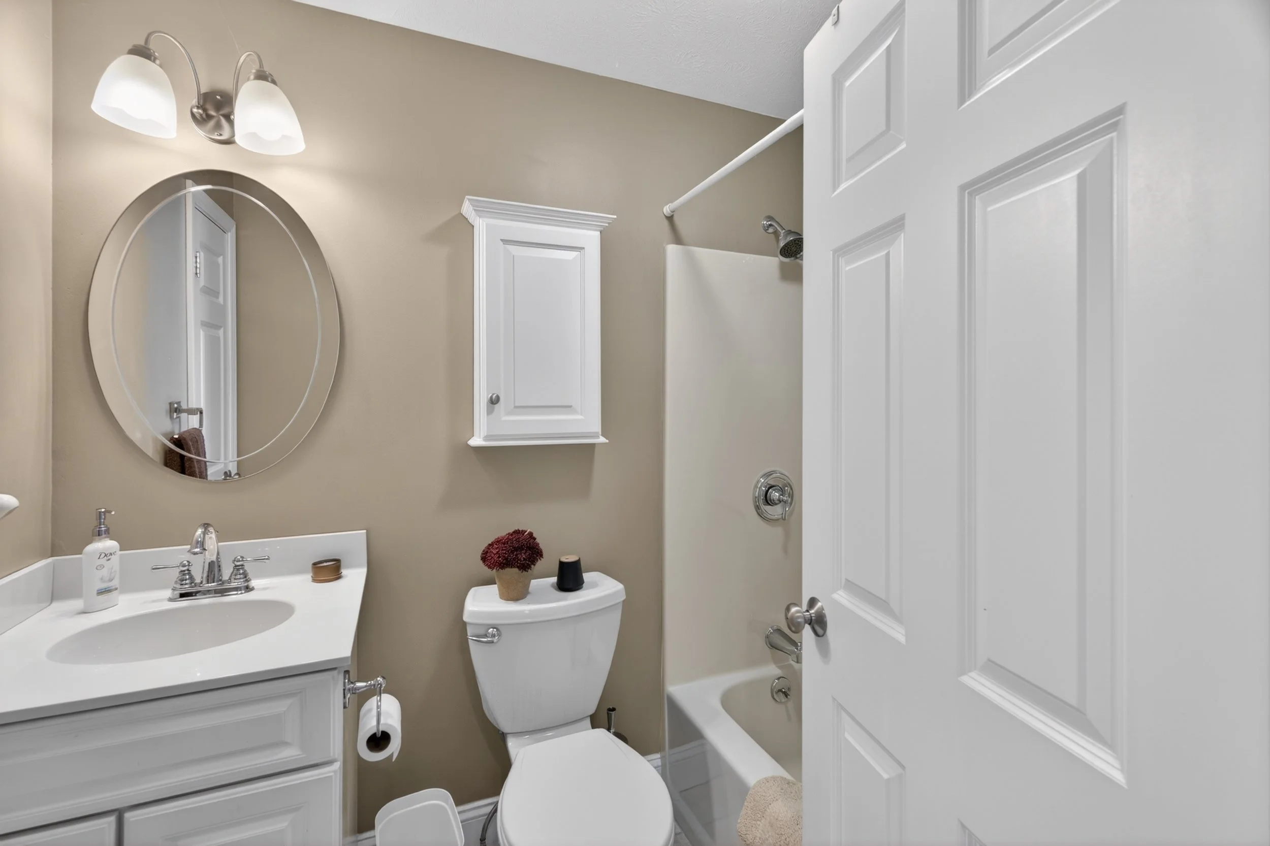 A small bathroom with beige walls and white fixtures. It includes a white vanity with a silver faucet, a round mirror, a white toilet with a flower pot and other small decorations on top, a white cabinet on the wall, a bathtub with a showerhead, and 