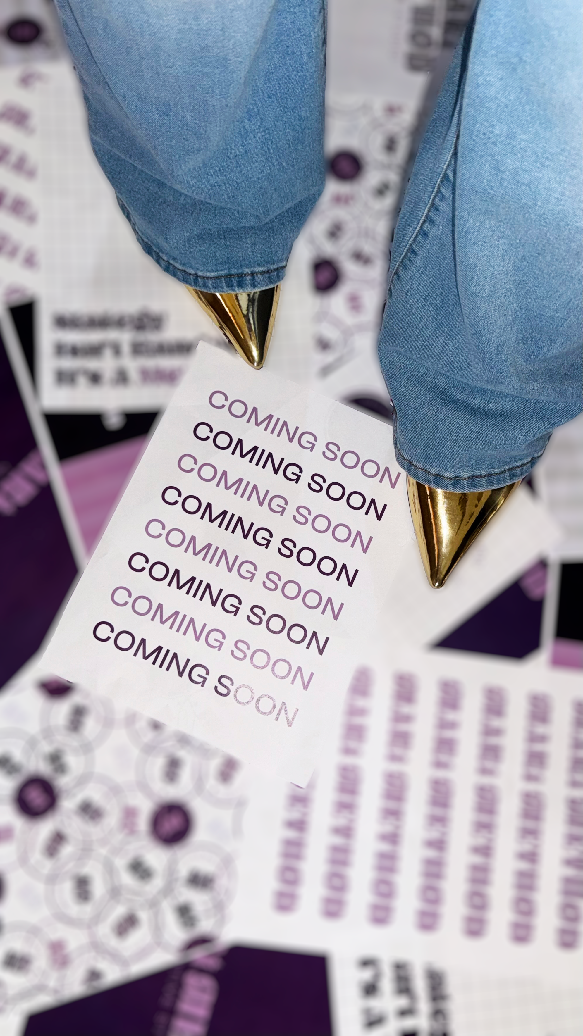 A close-up of a person's legs wearing blue jeans and gold-tipped shoes, standing on a surface with multiple 'Coming Soon' stacked paper.