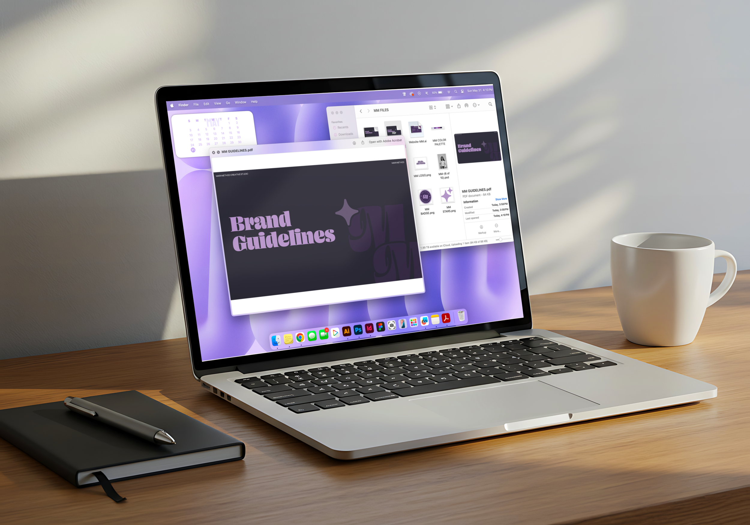 A laptop on a wooden desk displaying a graphic design project titled 'Brand Guidelines' with various folders and files open on the screen. A white coffee mug is situated to the right, close to the laptop. A black notebook and pen are on the left side of the desk, and the background features a gray wall with sunlight shadow patterns.