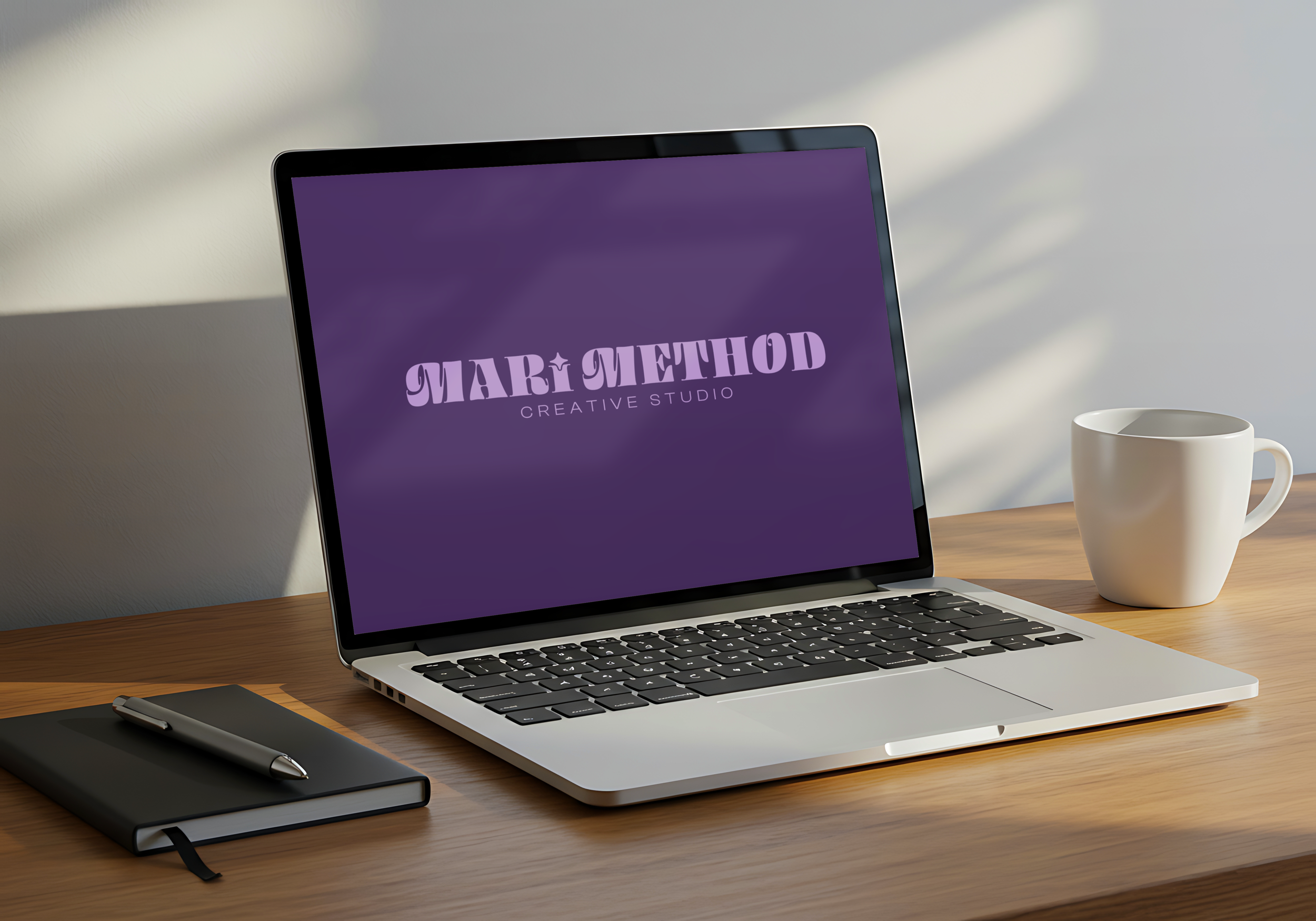 A laptop displaying the logo of a creative studio called 'MARI METHOD'. The laptop is on a wooden desk, accompanied by a closed notebook with a pen on top, a white coffee mug, and a gray smartphone.