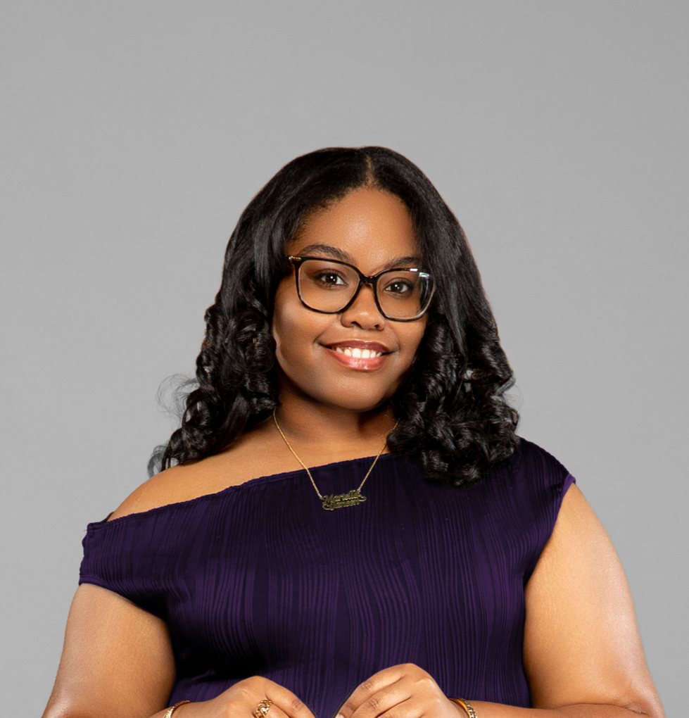 A woman with long black curly hair, glasses, and a purple off-shoulder top smiling for the camera against a gray background.