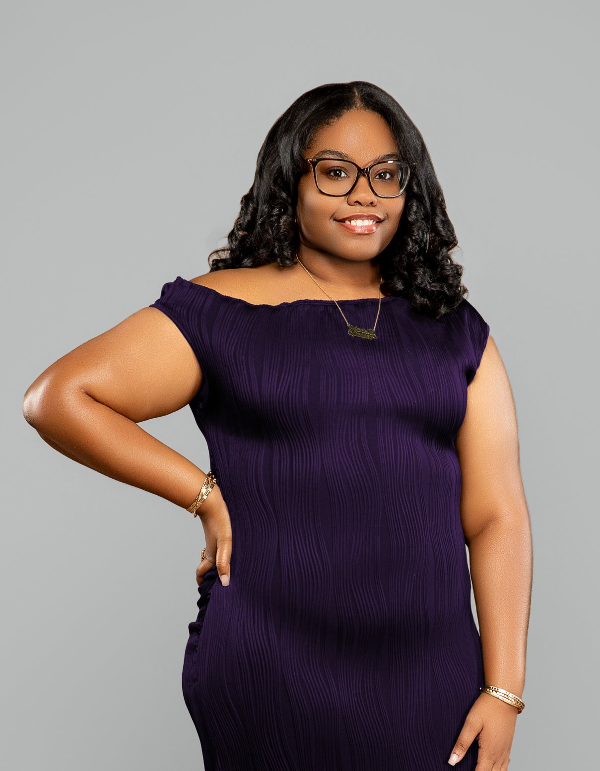A woman with dark, curly hair, wearing glasses, a purple dress, and gold jewelry, standing against a gray background.