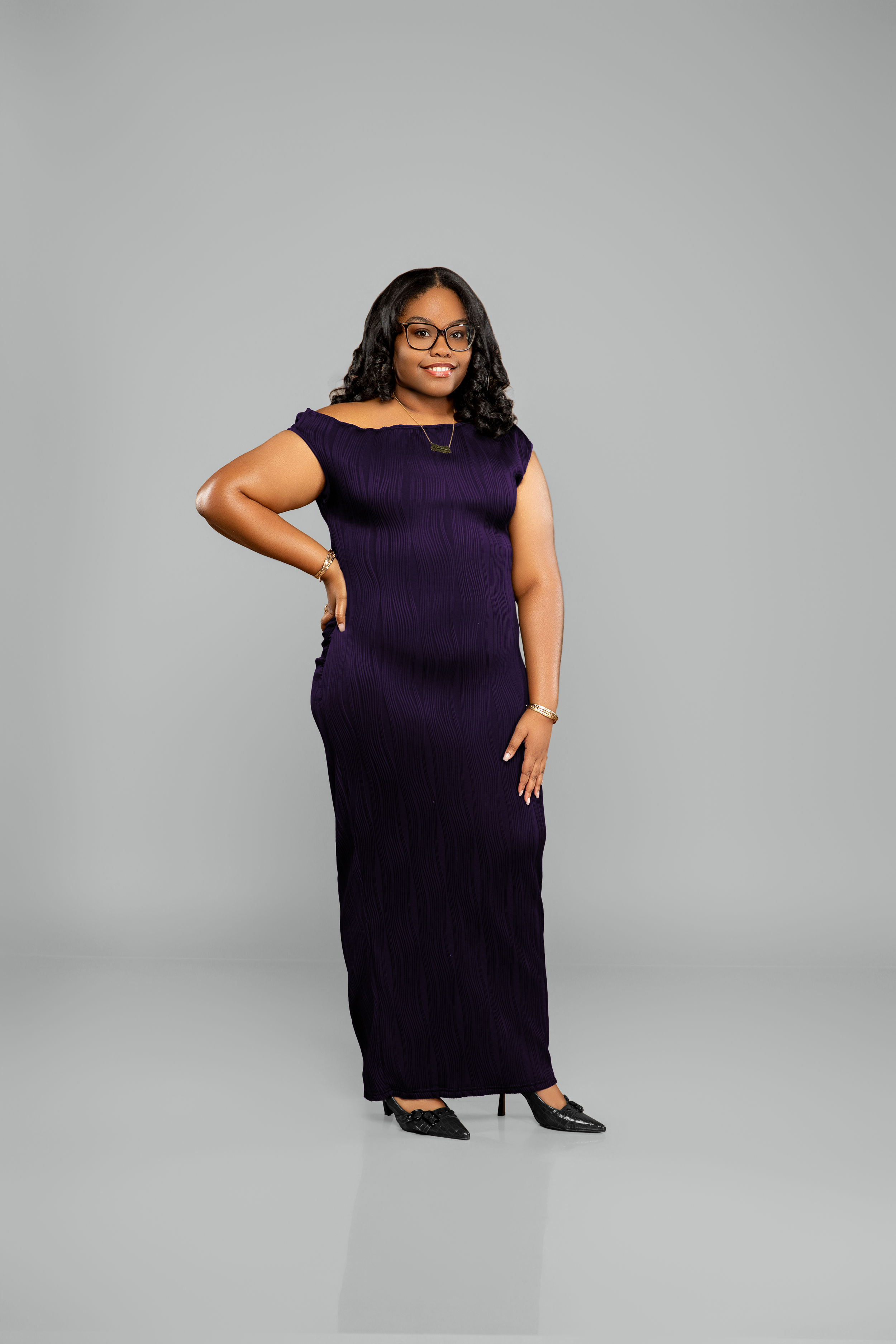 A woman with black hair, glasses, and a purple dress standing against a plain gray background, smiling with one hand on her hip and the other hanging by her side.