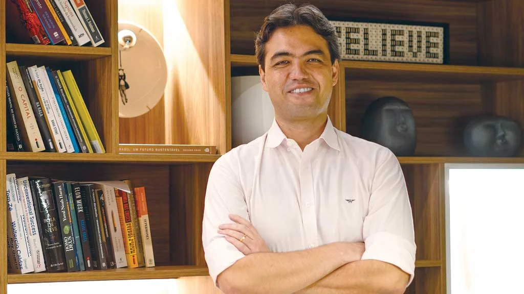 Homem sorridente de camisa branca cruzando os braços, em um ambiente com biblioteca de madeira ao fundo e objetos decorativos.