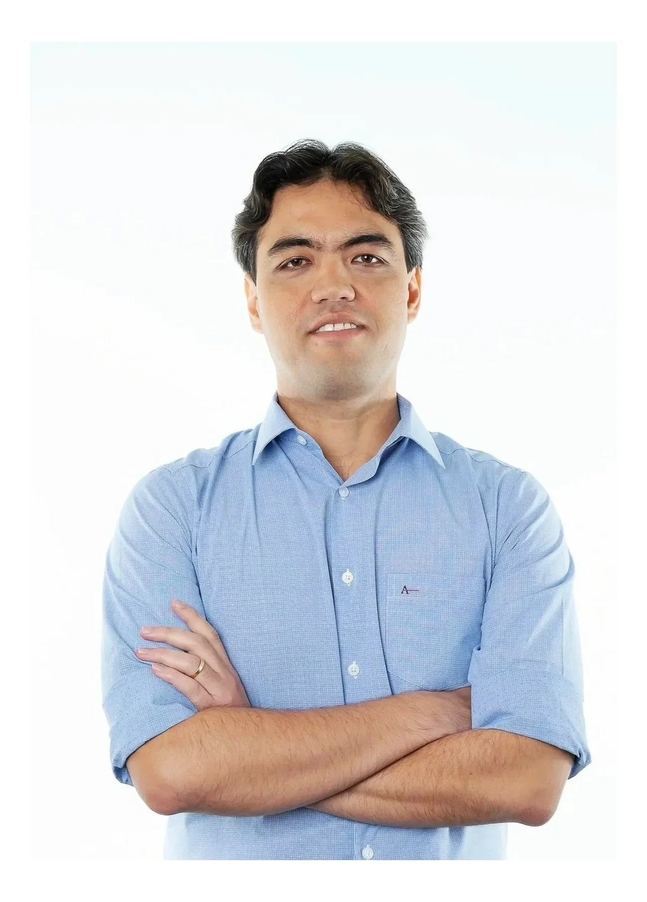 Homem de cabelo escuro e pele clara, vestindo uma camisa social azul, com os braços cruzados, em fundo branco