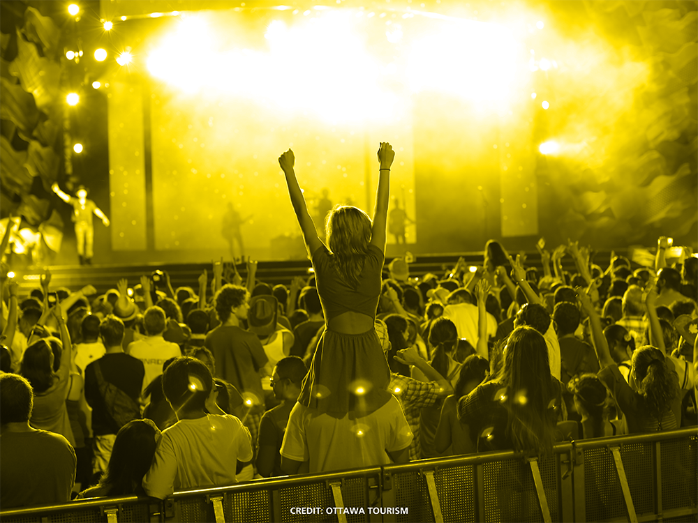 image of a young woman raising her arms in a crowd at a concert. A yellow glow illuminates the crowd and musicians.