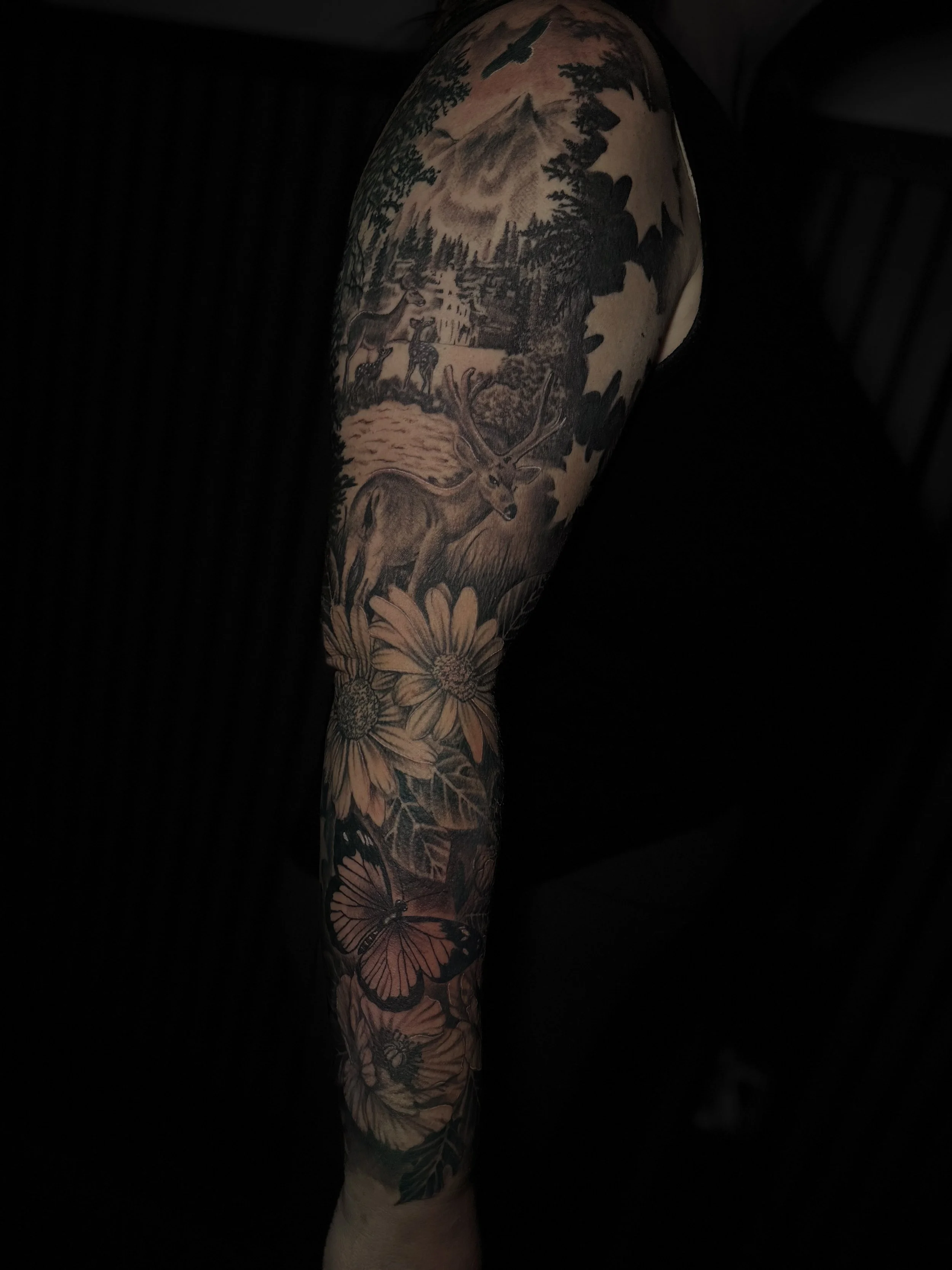 Tattoo sleeve depicting a nature scene with mountains, trees, a river, a deer with large antlers, and flowers.