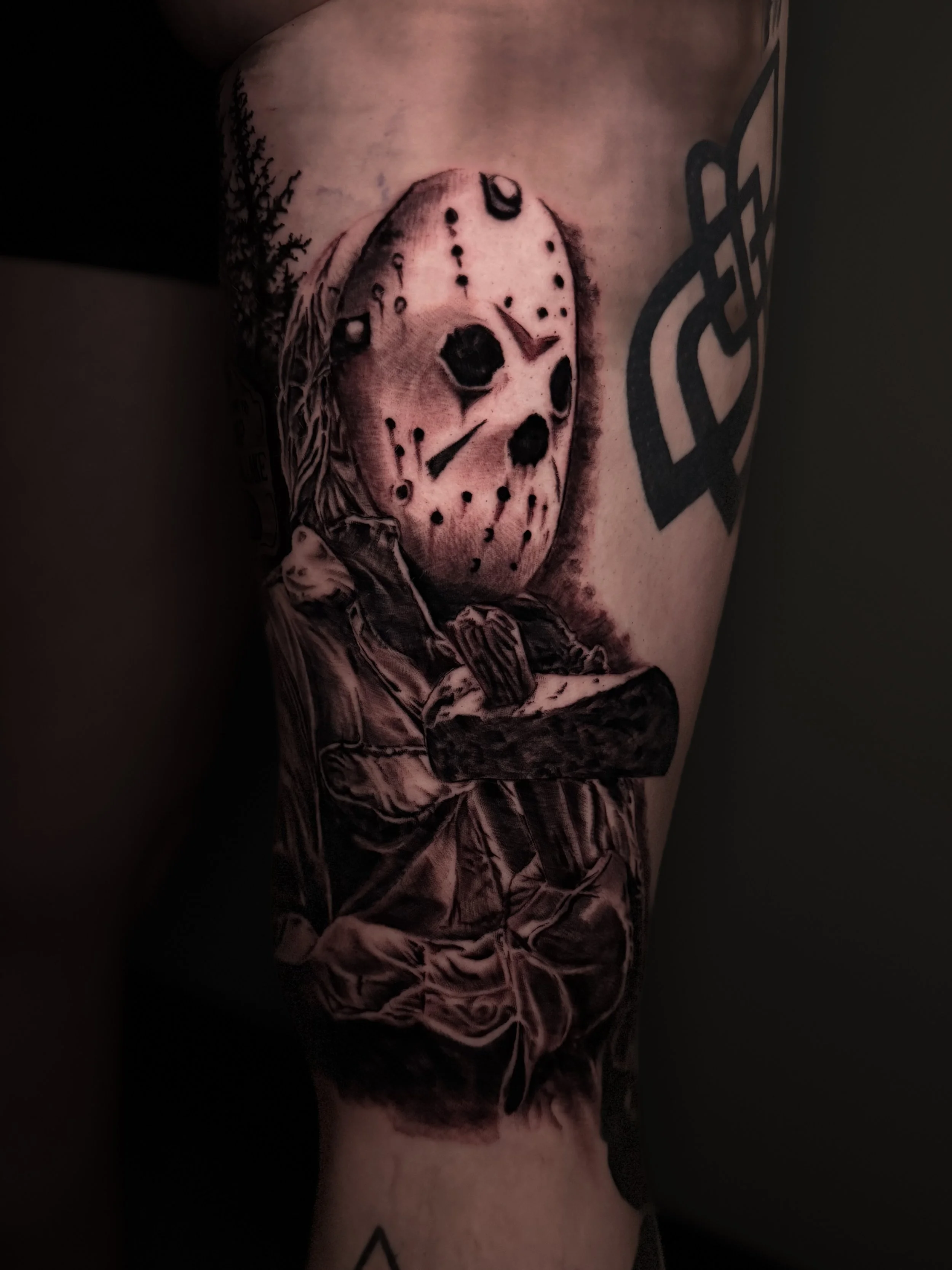 Tattoo of a person in a hockey mask from Friday the 13th, with a forest background on the shoulder, and a geometric black tattoo on the arm.