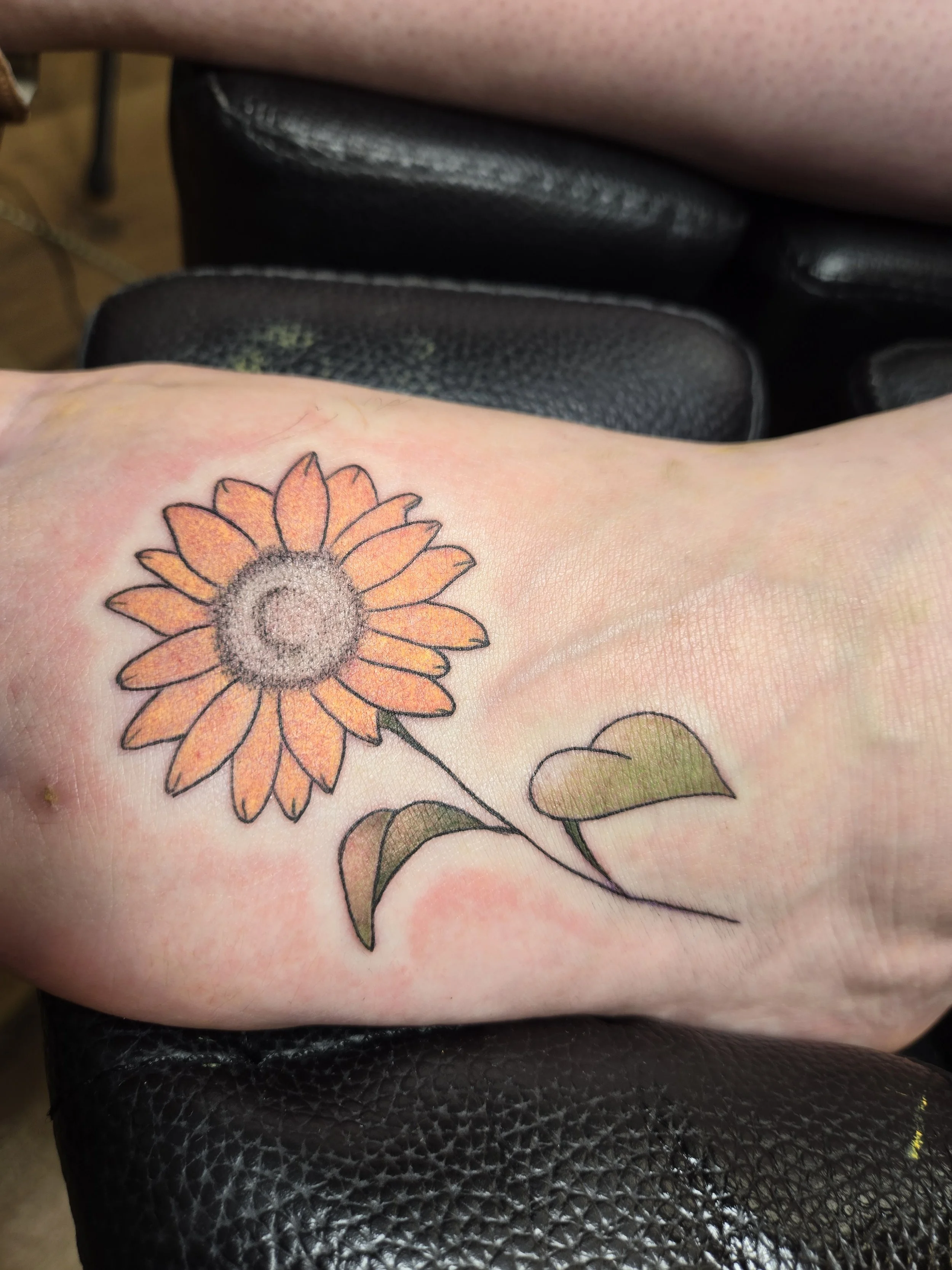 A person's arm with a sunflower tattoo and two green leaves on the skin, resting on a black textured surface, with part of another person's leg in the background.