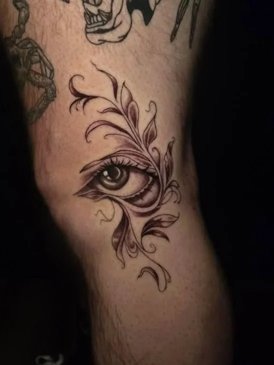 Tattoo of a realistic eye with ornamental leaves and swirls on forearm.