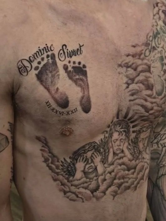 Tattoo of two baby footprints with the words 'Dominic Swet' and Roman numerals 'XII-XIX-XX' above the footprints, and a tattoo of a solemn angel surrounded by clouds and rays on the person's chest.