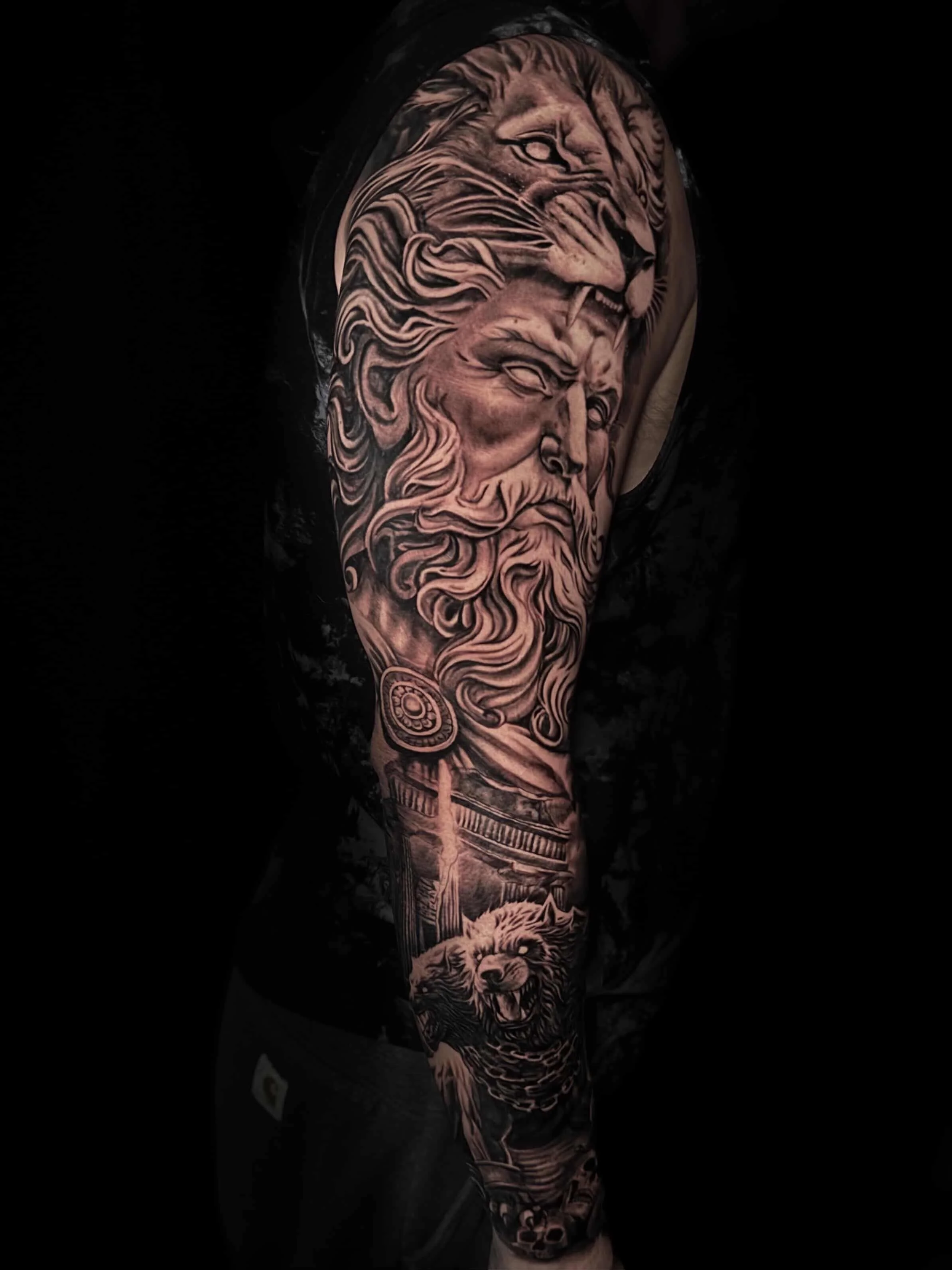 Tattooed arm featuring a detailed black and gray tattoo of a lion's face and mane, with elements like a circular design and a lion's head at the bottom.