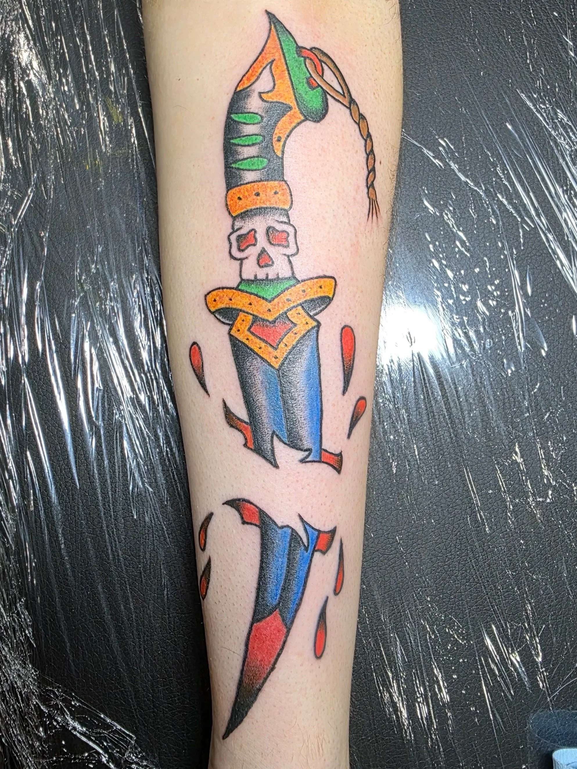 Colorful tattoo of a dagger with a skull in the middle, decorated with orange, green, and black details, on a person's forearm.