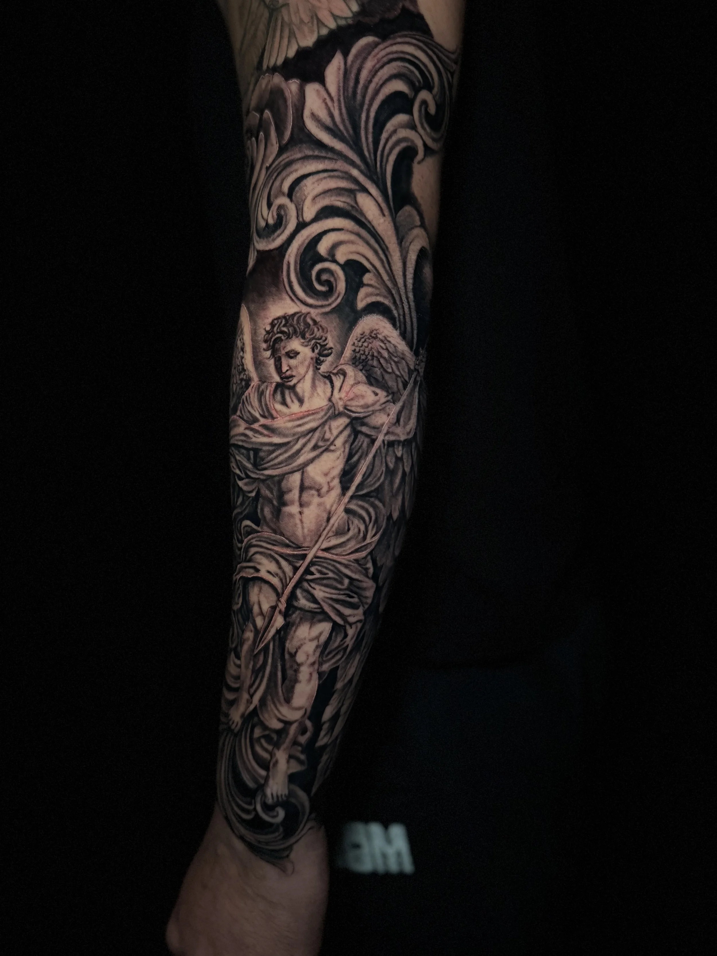 Tattoo of an angel with wings and a flowing robe, surrounded by swirling patterns, on a person's forearm.