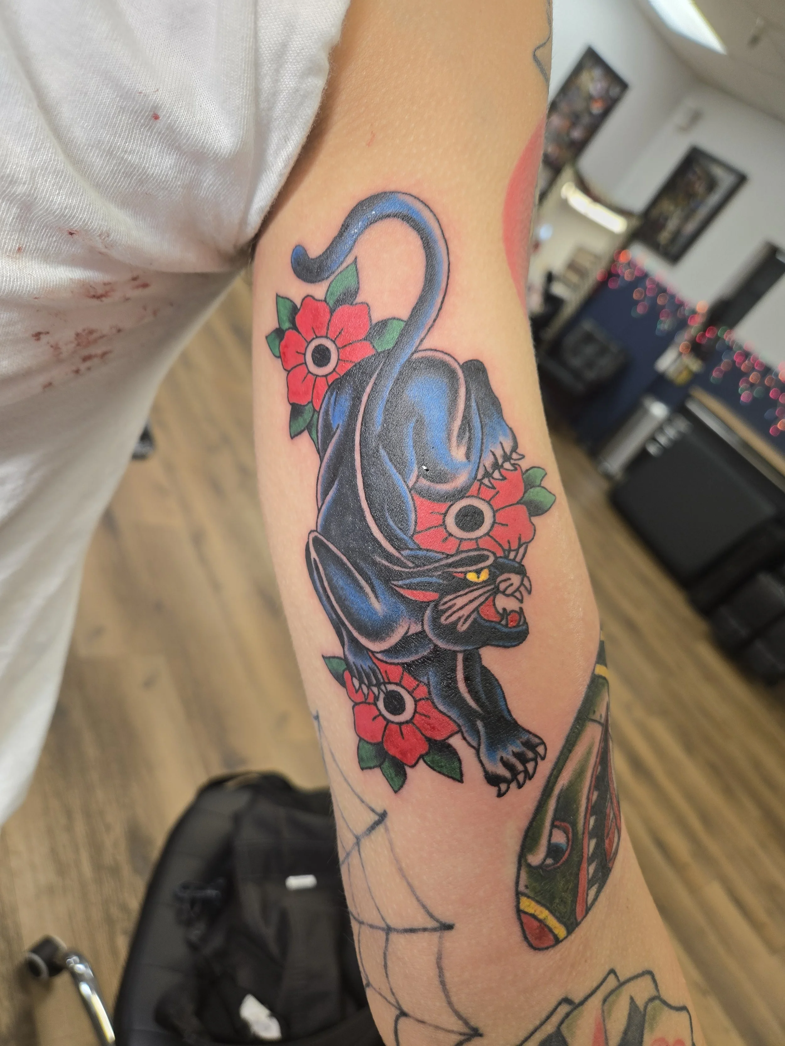 A colorful tattoo of a black cat with yellow eyes lying down, surrounded by red flowers and green leaves, on a person's arm.