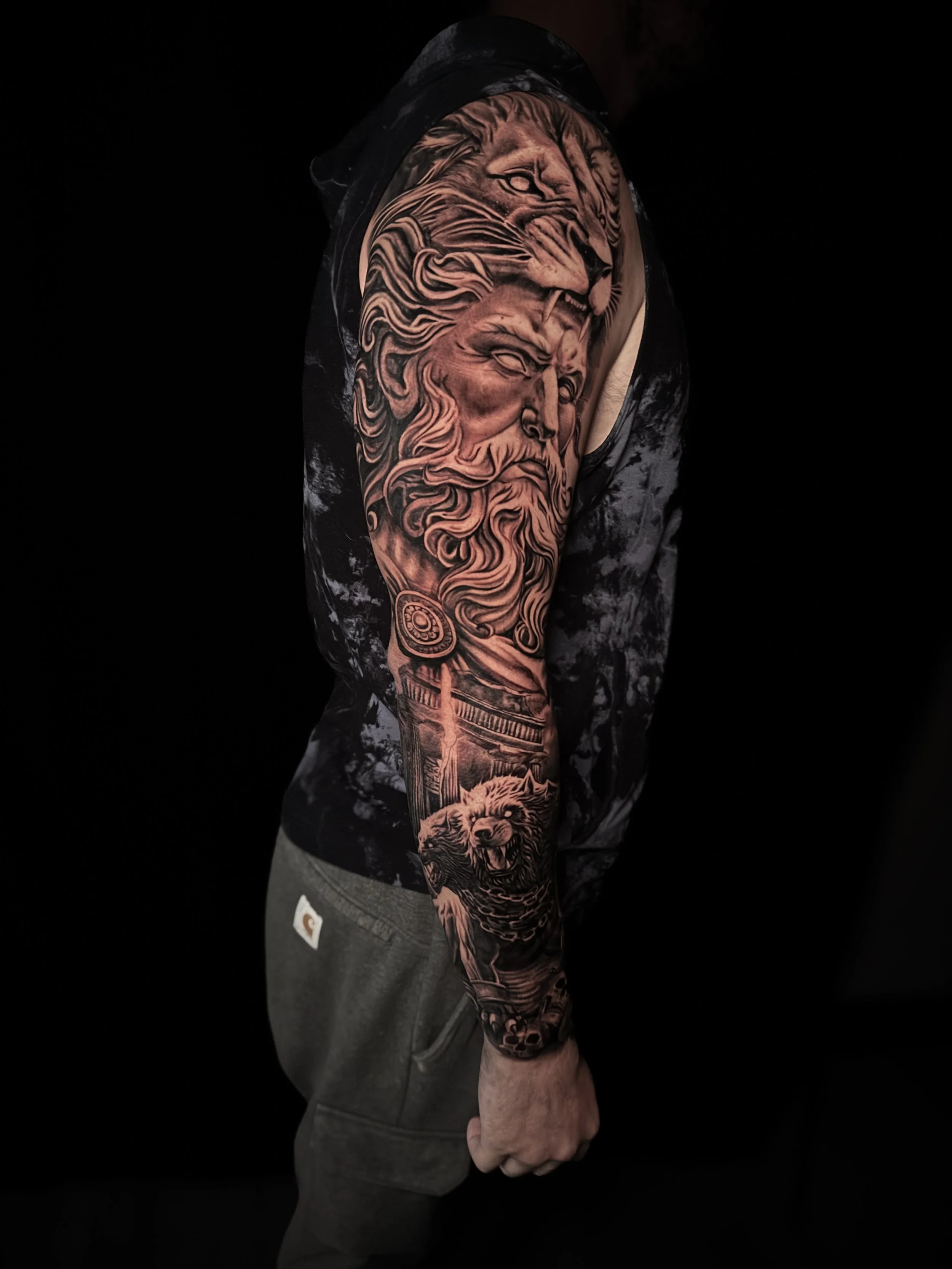 A person with a sleeve tattoo featuring a lion and a mythological creature on their arm, standing against a black background.
