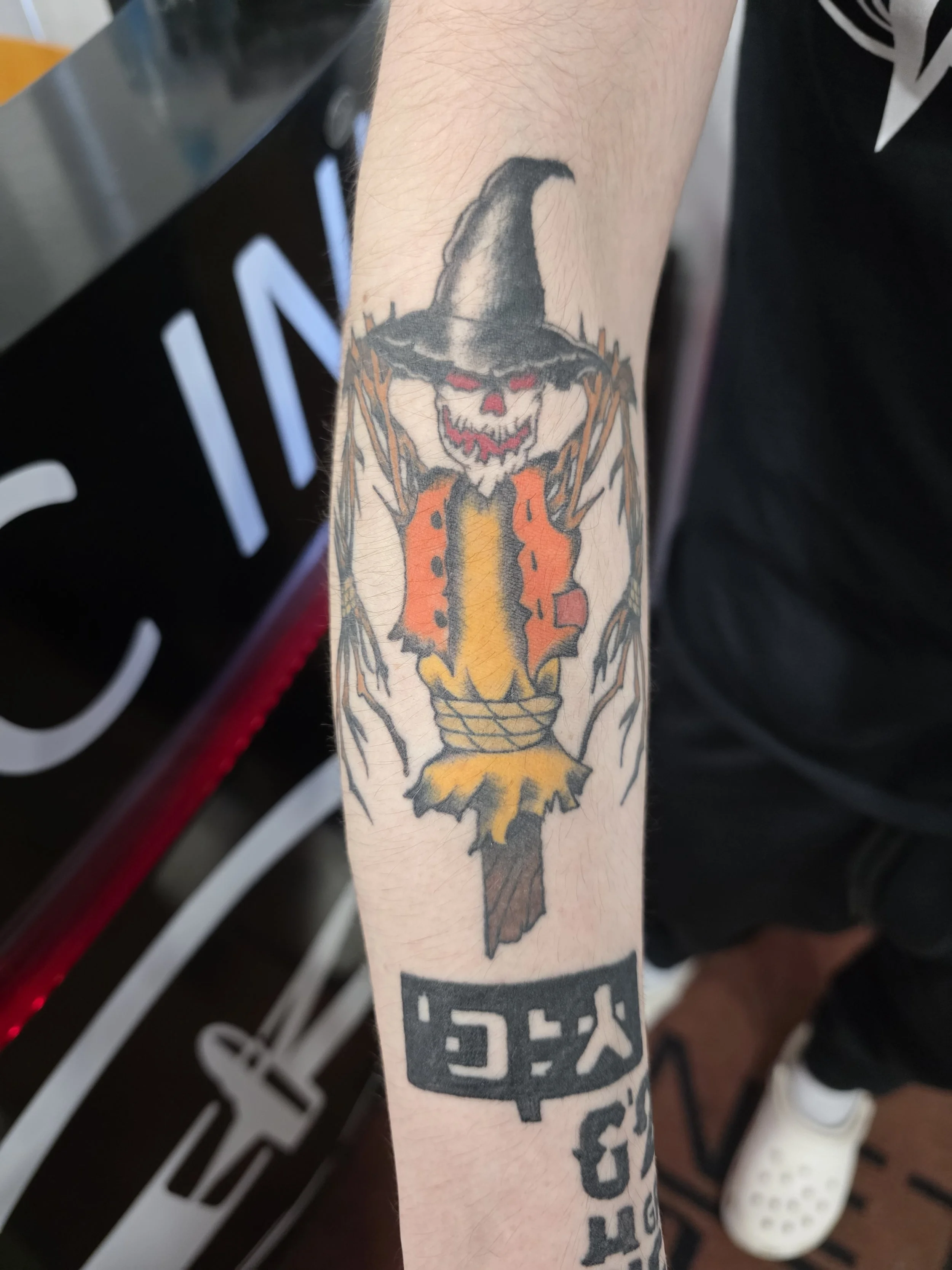 Tattoo of a creepy clown with red eyes, wearing a black witch hat, in front of a yellow flame, on a person's arm.