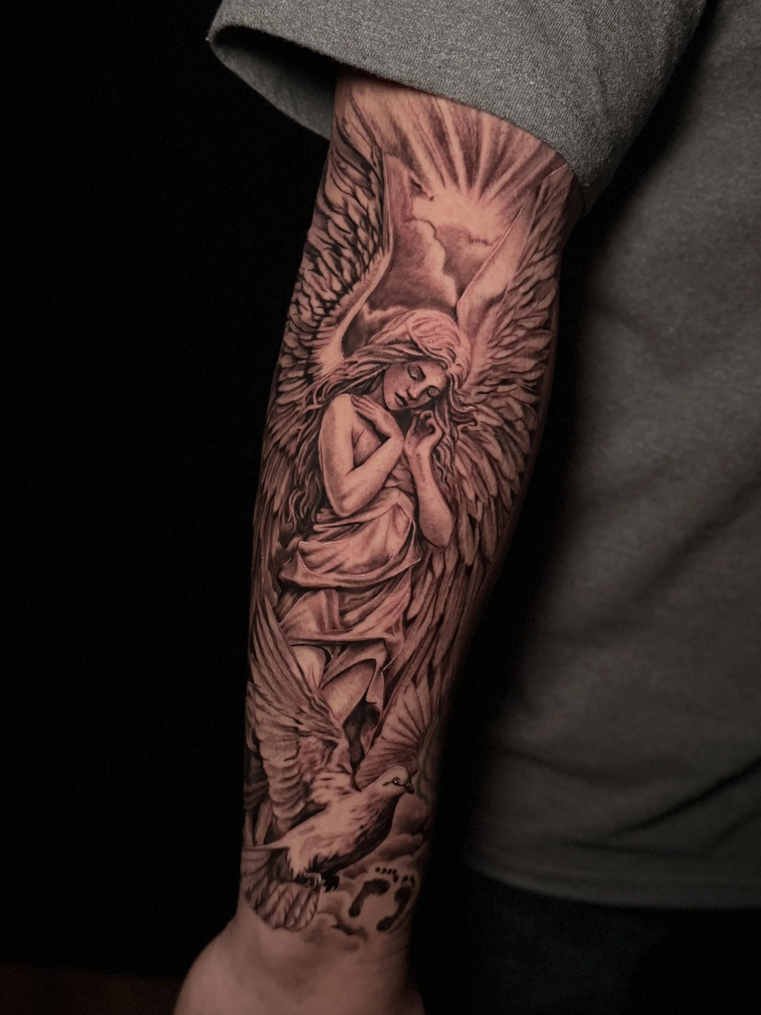 A tattoo sleeve featuring a detailed black and gray portrait of an angel with large wings and a peaceful expression, covering the entire arm from shoulder to wrist.