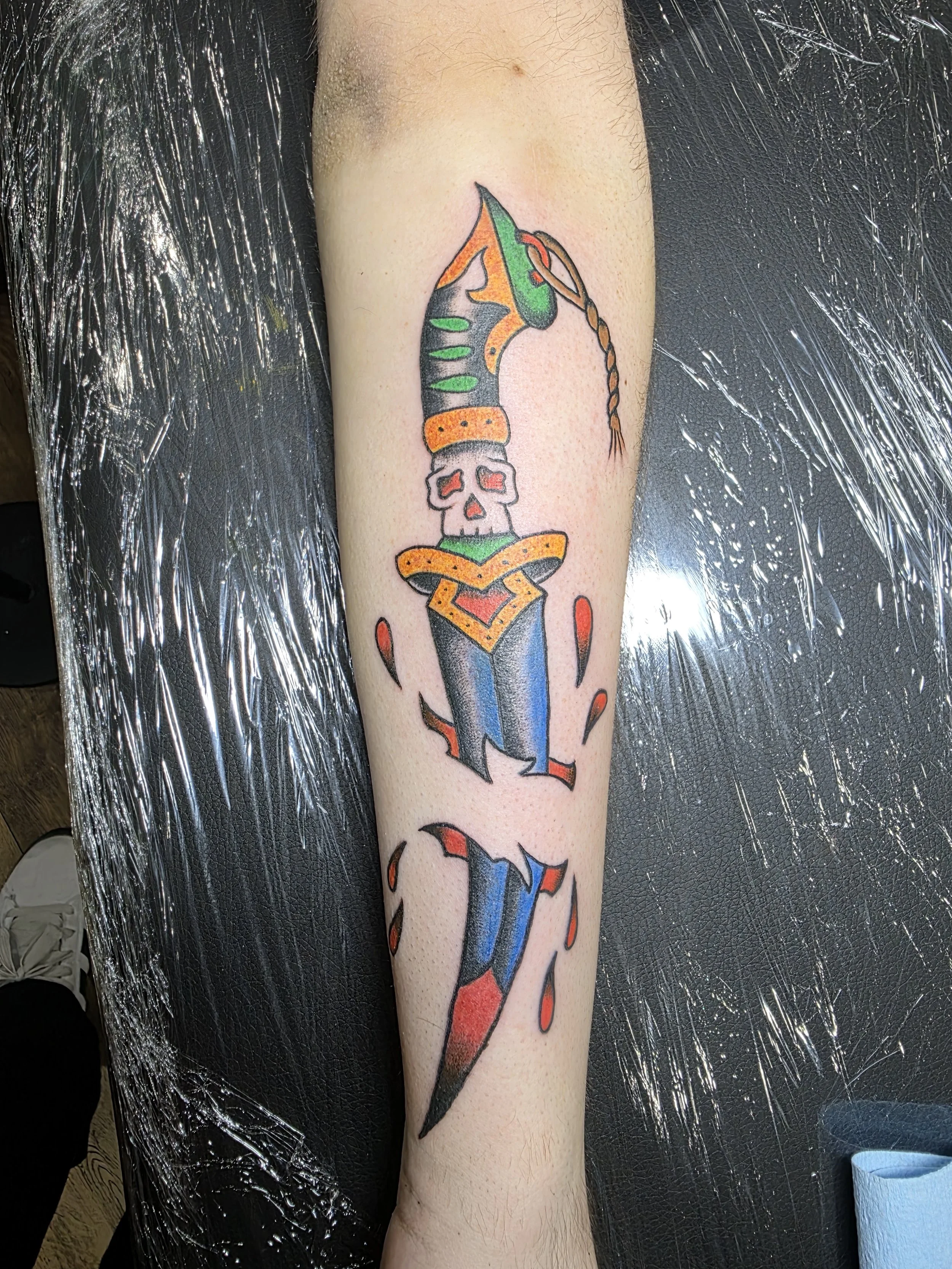 Colorful tattoo on person's arm featuring a skull with a dagger through it, decorated with orange, green, blue, and black ink.