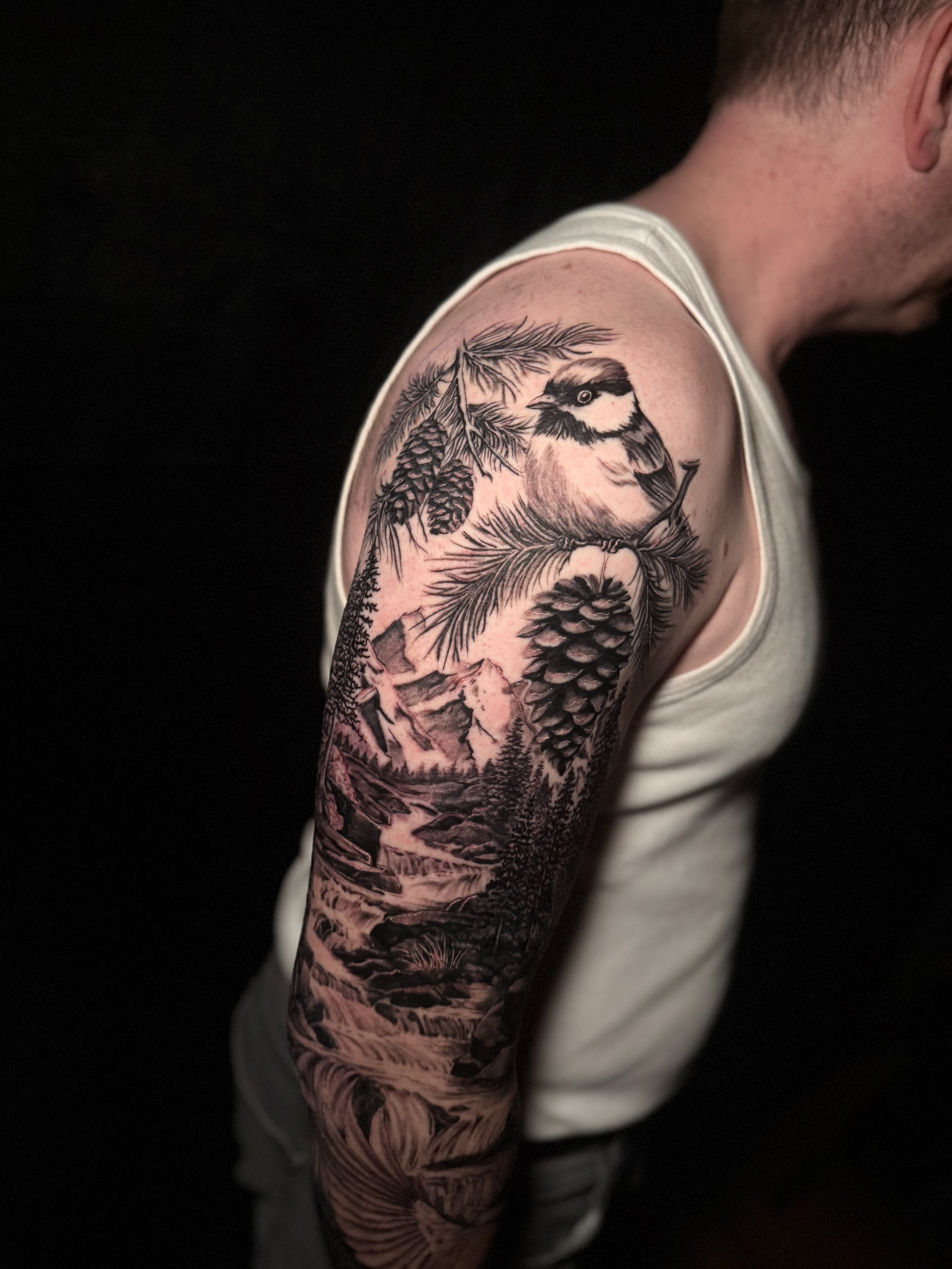 Black and gray tattoo of a bird on a pine branch on man's upper arm.