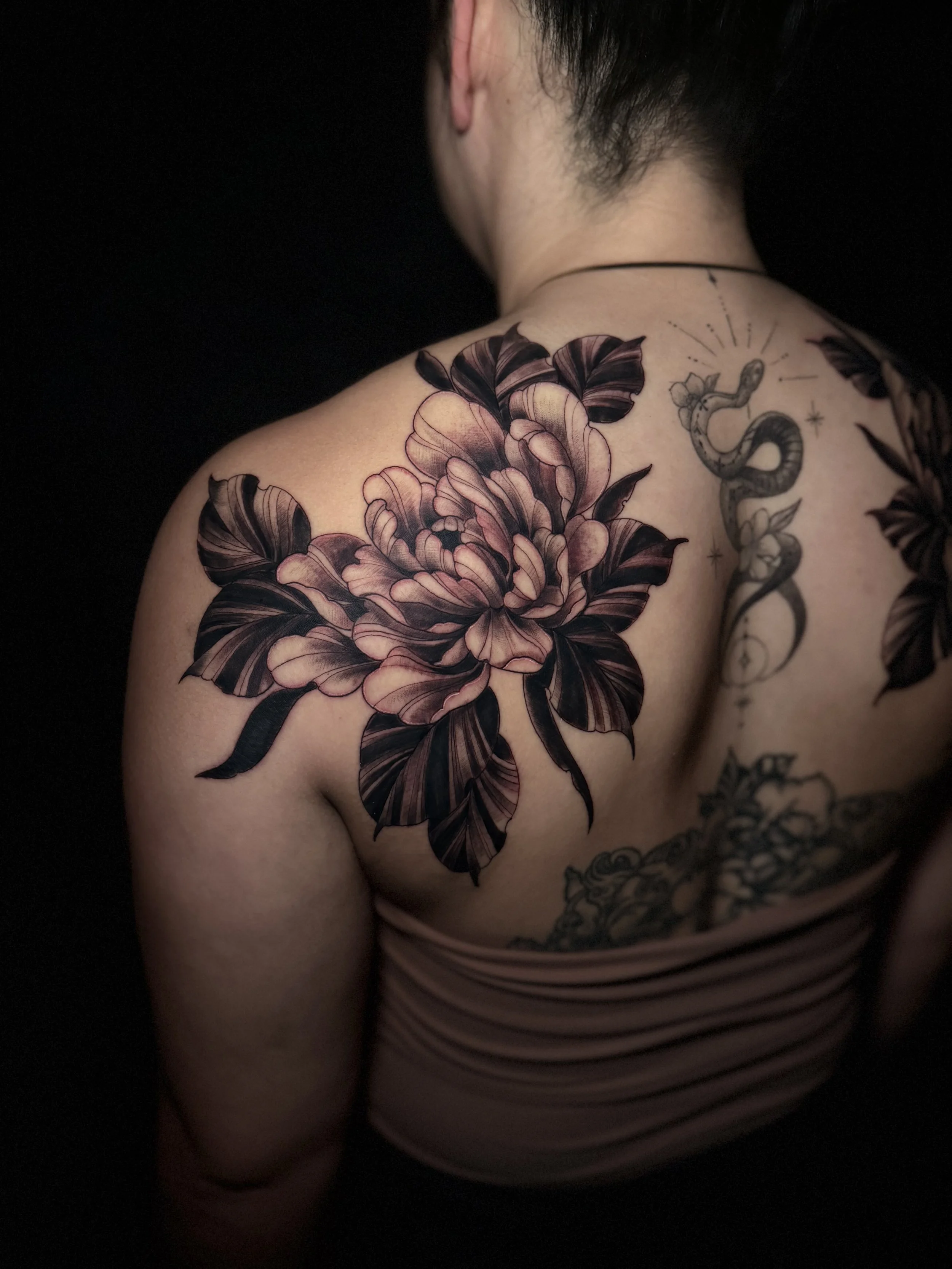 Tattooed shoulder of a person with various black and grey tattoos, including a large flower and a snake, against a dark background.