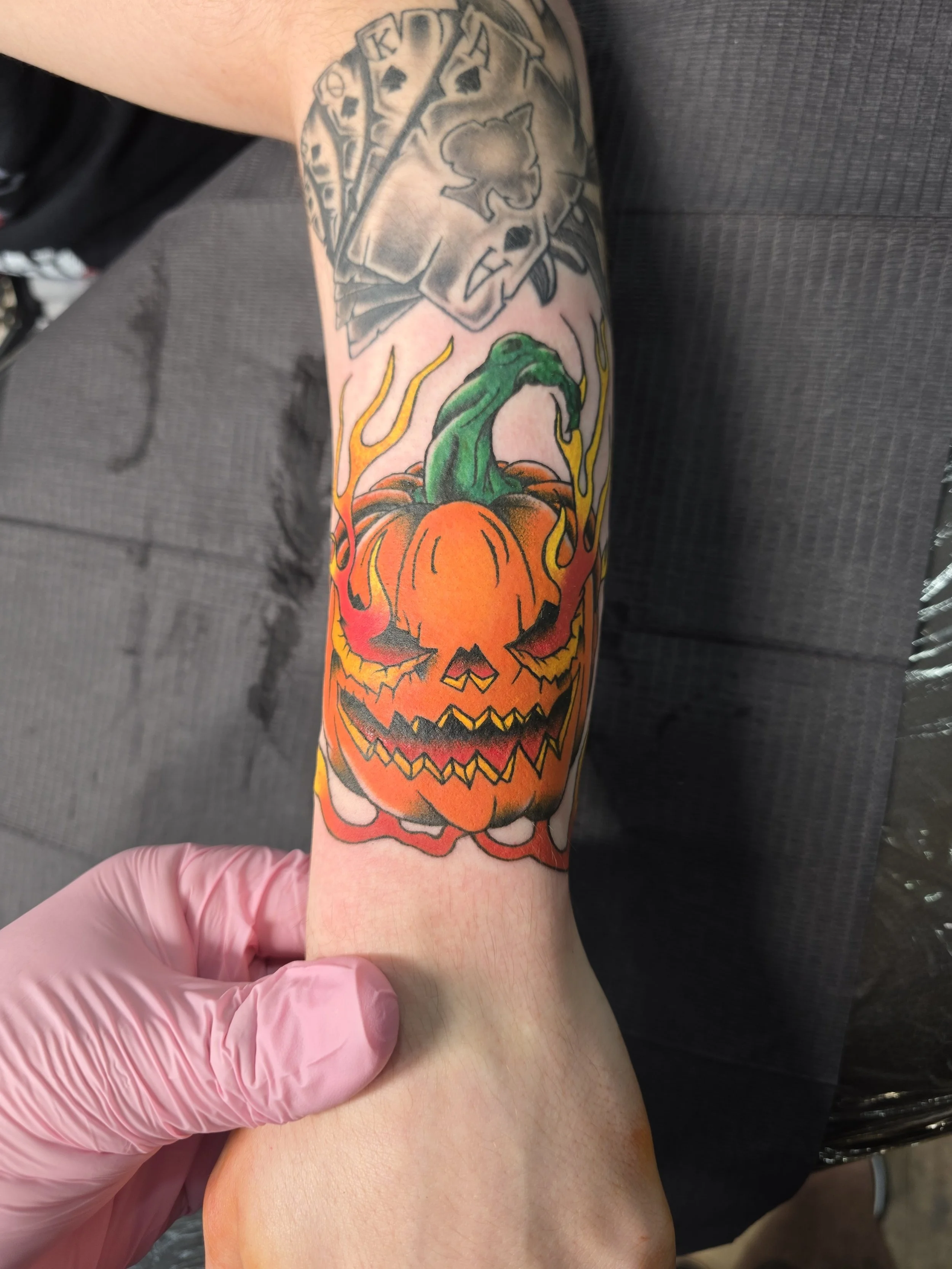 A colorful tattoo featuring a grinning jack-o'-lantern with a green, zombie-like creature emerging from inside, surrounded by flames, on a person's forearm.