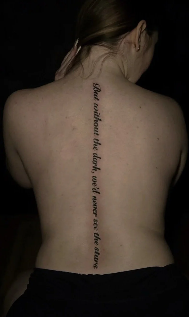 A person's back showing a vertical tattoo with the text "But without the dark, we'd never see the stars."